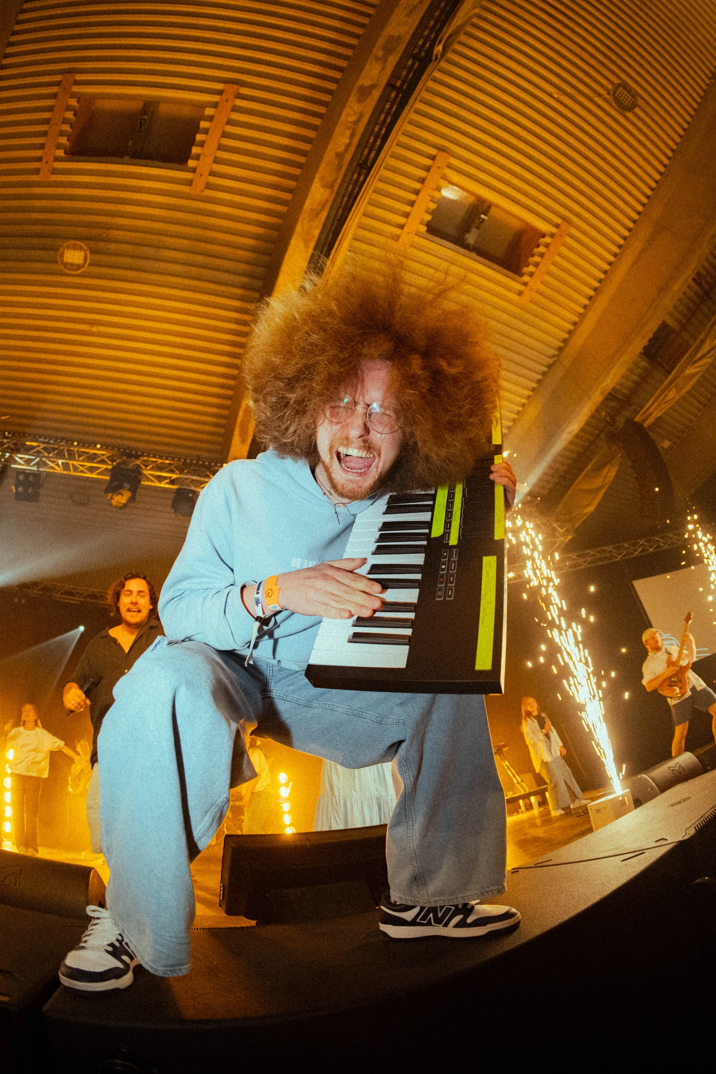 En person med stor, voluminøs hår sitter og spiller et keyboard på en scene med lys og folk rundt, og ser ut til å ha det gøy.