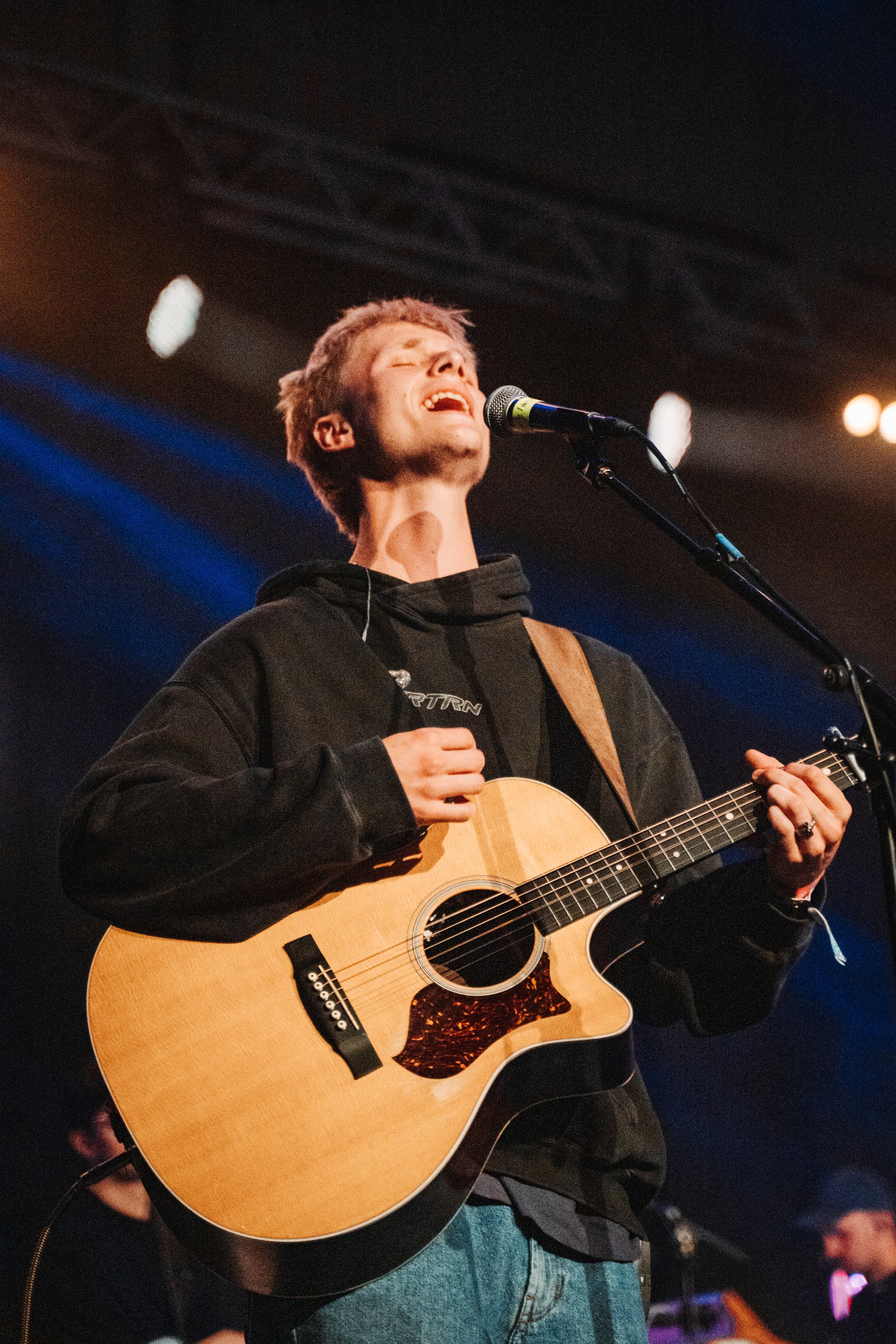 En mann spiller akustisk gitar på en scene med mørk bakgrunn og lys. Han har kort, lett støl hår og er iført svart hettegenser.