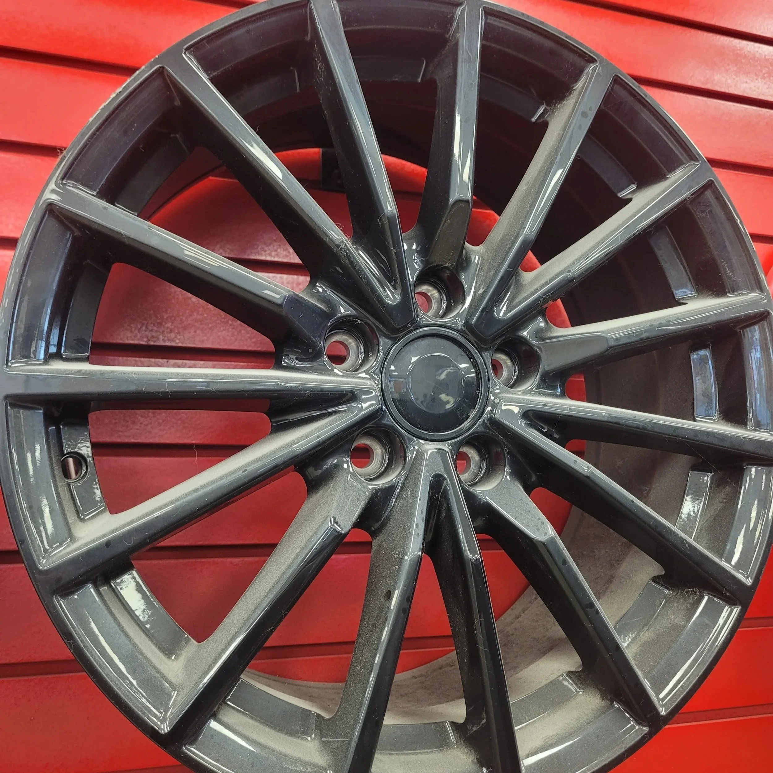 Deux jantes en alliage automobile, noires avec reflets, posées sur un fond rouge à rainures horizontales.
