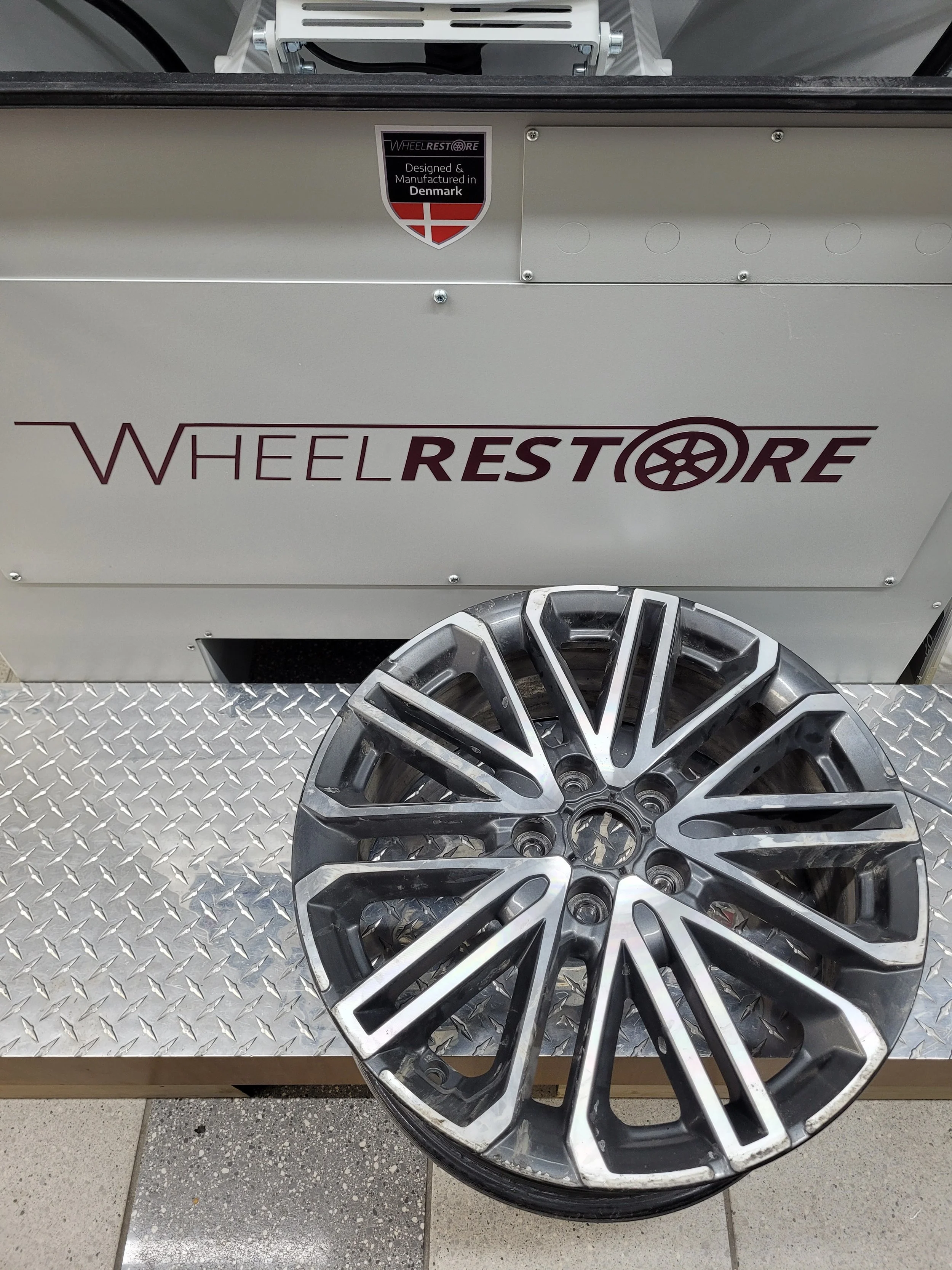 Une jante de voiture noire et argentée posée sur un sol en carrelage, avec un panneau en métal derrière avec le logo 'Wheelrestore' et une pièce de mobilier blanc.