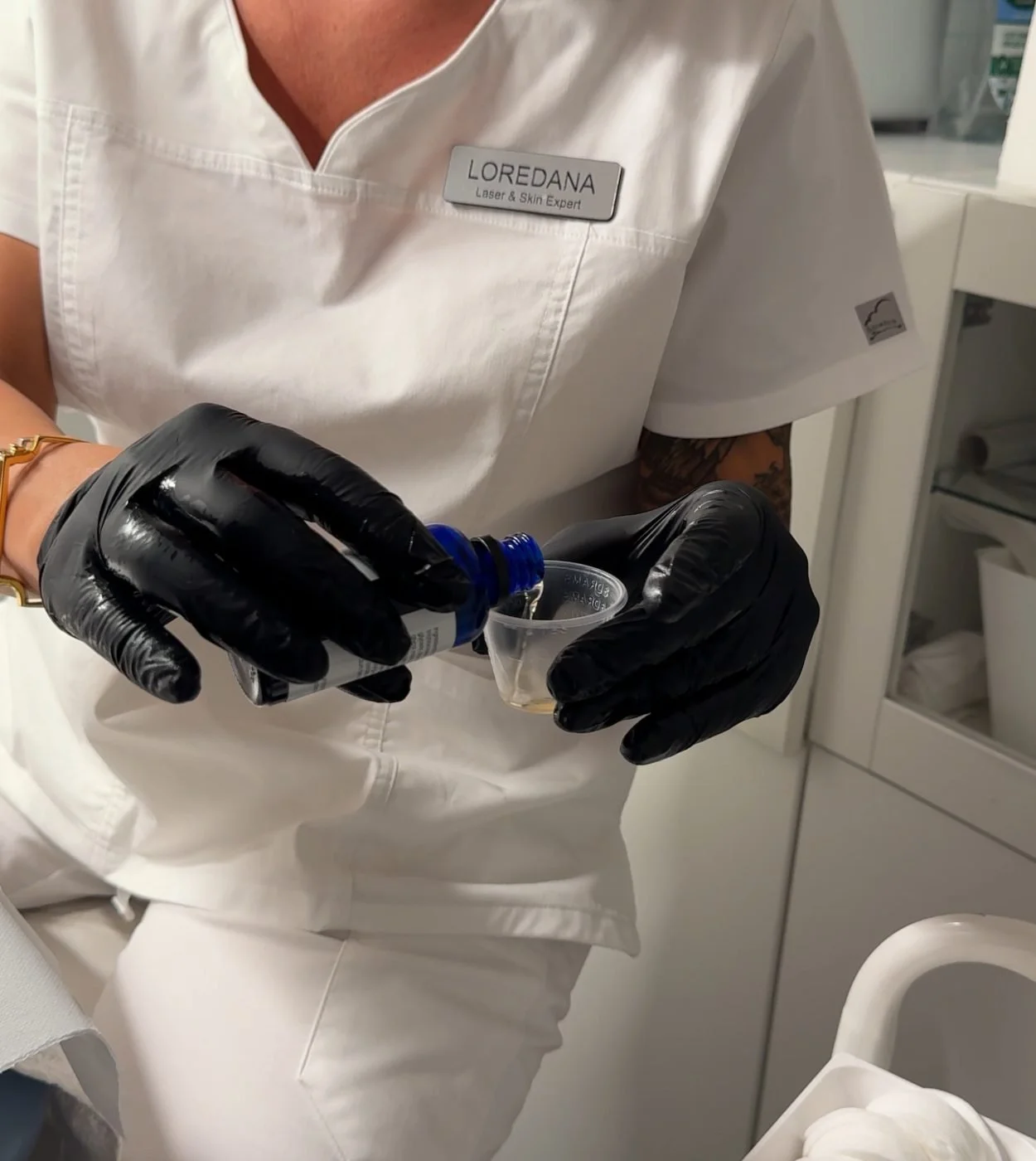 A healthcare professional wearing black gloves, a white uniform with a name tag 'LOREDANA', and a tattoo on the arm, is pouring a liquid into a small clear cup in a medical setting.