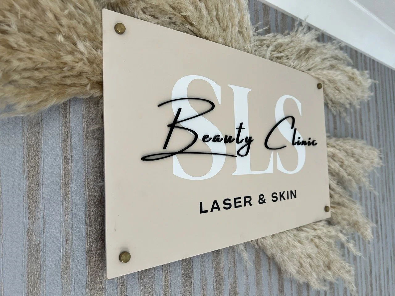 Sign for Belle Beauty Clinic with text "BELLE Beauty Clinic LASER & SKIN" on beige background, mounted on wall with decorative pampas grass
