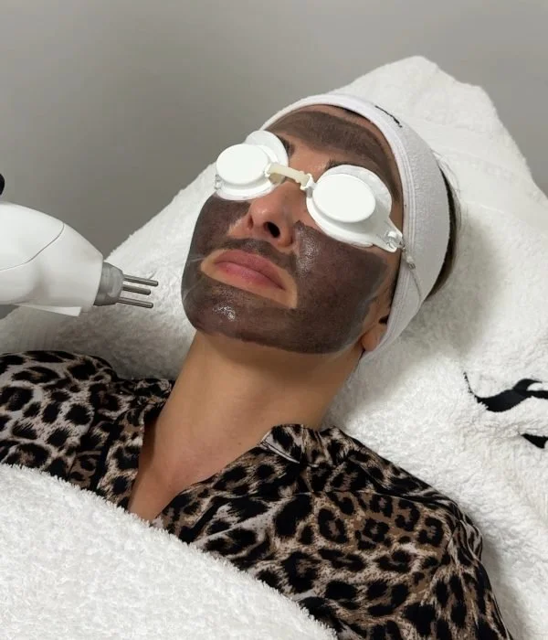 Woman with a dark facial mask, lying down with protective goggles and a headband, receiving facial treatment at a spa.
