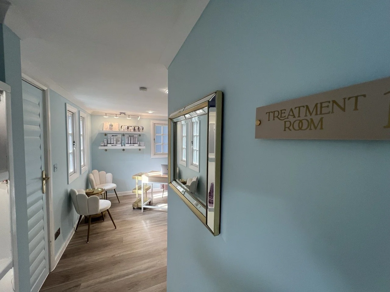 Interior view of a treatment room with light blue walls, white chairs, a small table, and shelves with medical supplies. A mirror is mounted on the wall, and sunlight flows through multiple windows.