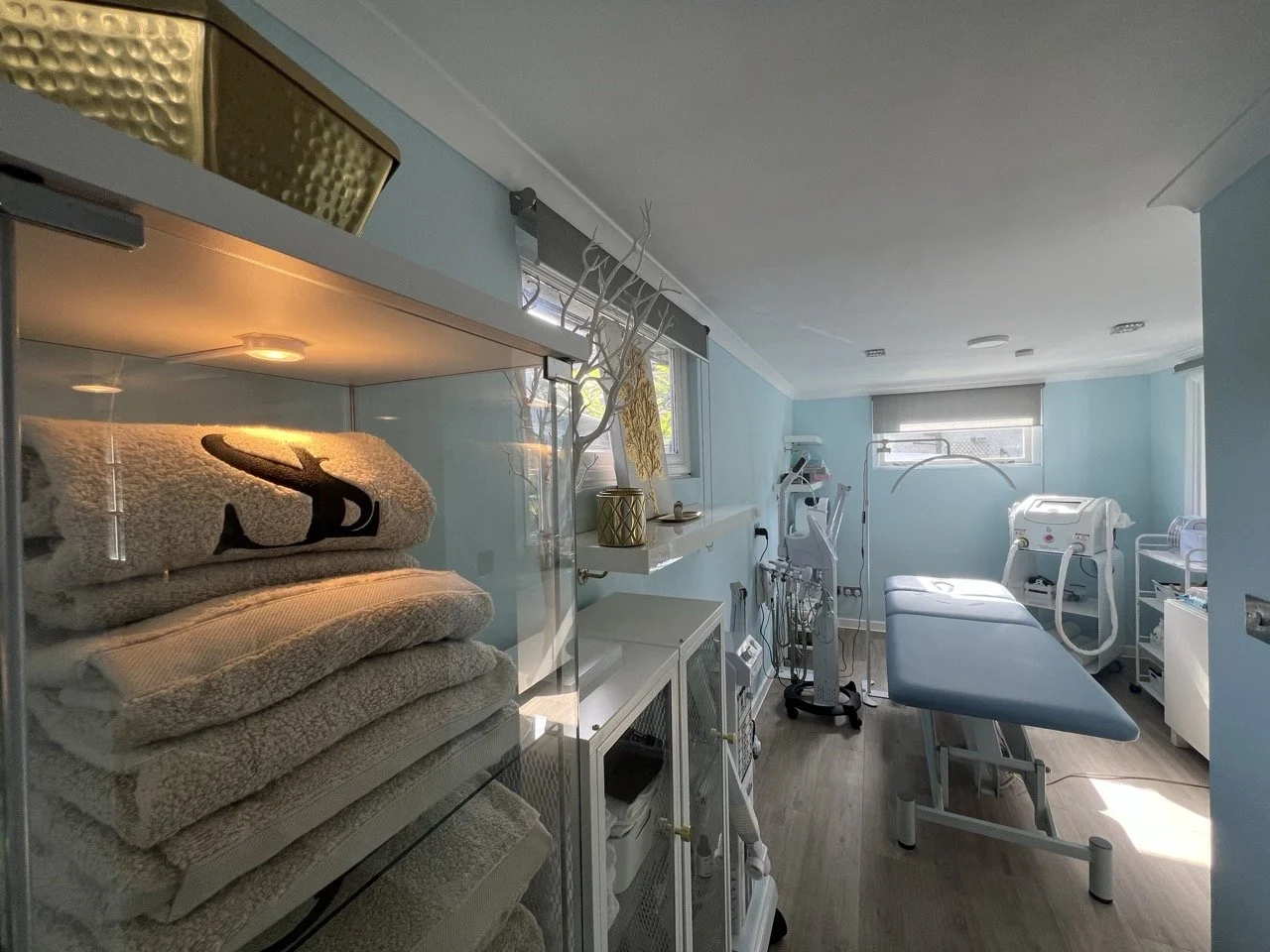 A medical examination room with a blue interior, including an examination table, medical equipment, and sunlight coming through the windows.