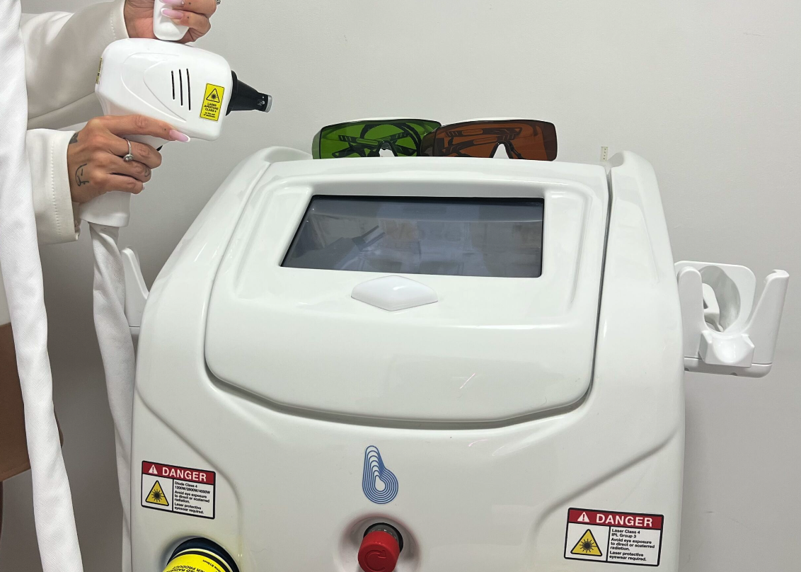 A person holds a handheld laser device aimed at a white machine with a screen, which has a transparent window, two pairs of sunglasses on top, and warning labels about laser dangers.