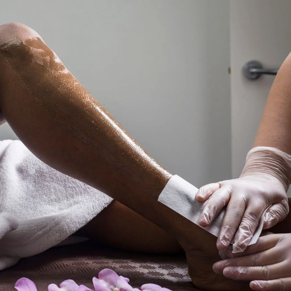 A person receiving a waxing treatment on their leg, with a technician applying wax and wearing gloves.