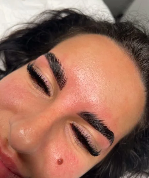 Close-up of a woman with long, thick eyebrows, eyelash extensions, and smooth skin, lying down with her eyes closed.