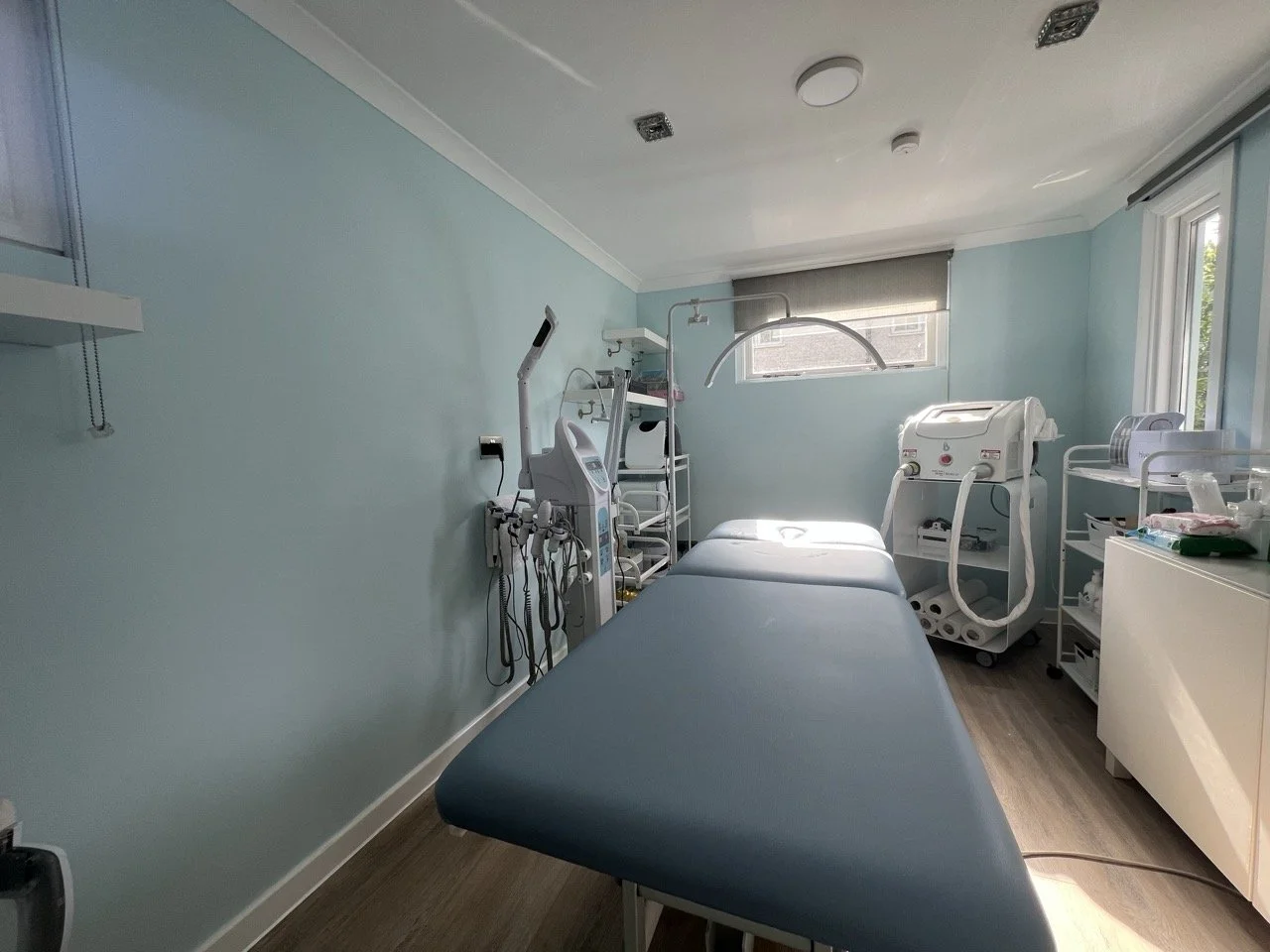 Empty medical examination room with a blue examination table, medical equipment, and supplies, sunlight coming through windows.