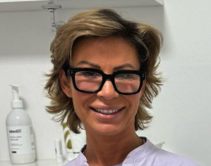 A woman with short blonde hair and glasses smiling in a medical or clinical setting, with white walls and medical supplies in the background.
