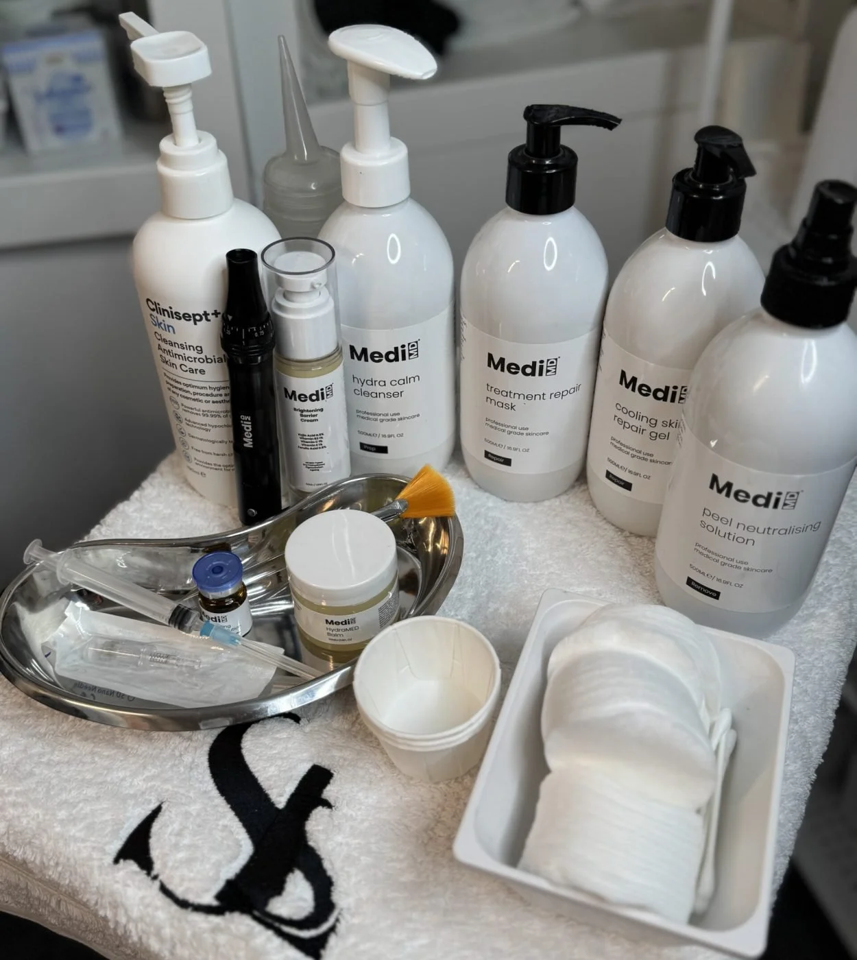 Various skincare and medical supplies on a table, including bottles of cleanser, mask, gel, solution, a small jar, a syringe, cotton pads, and disposable cups.