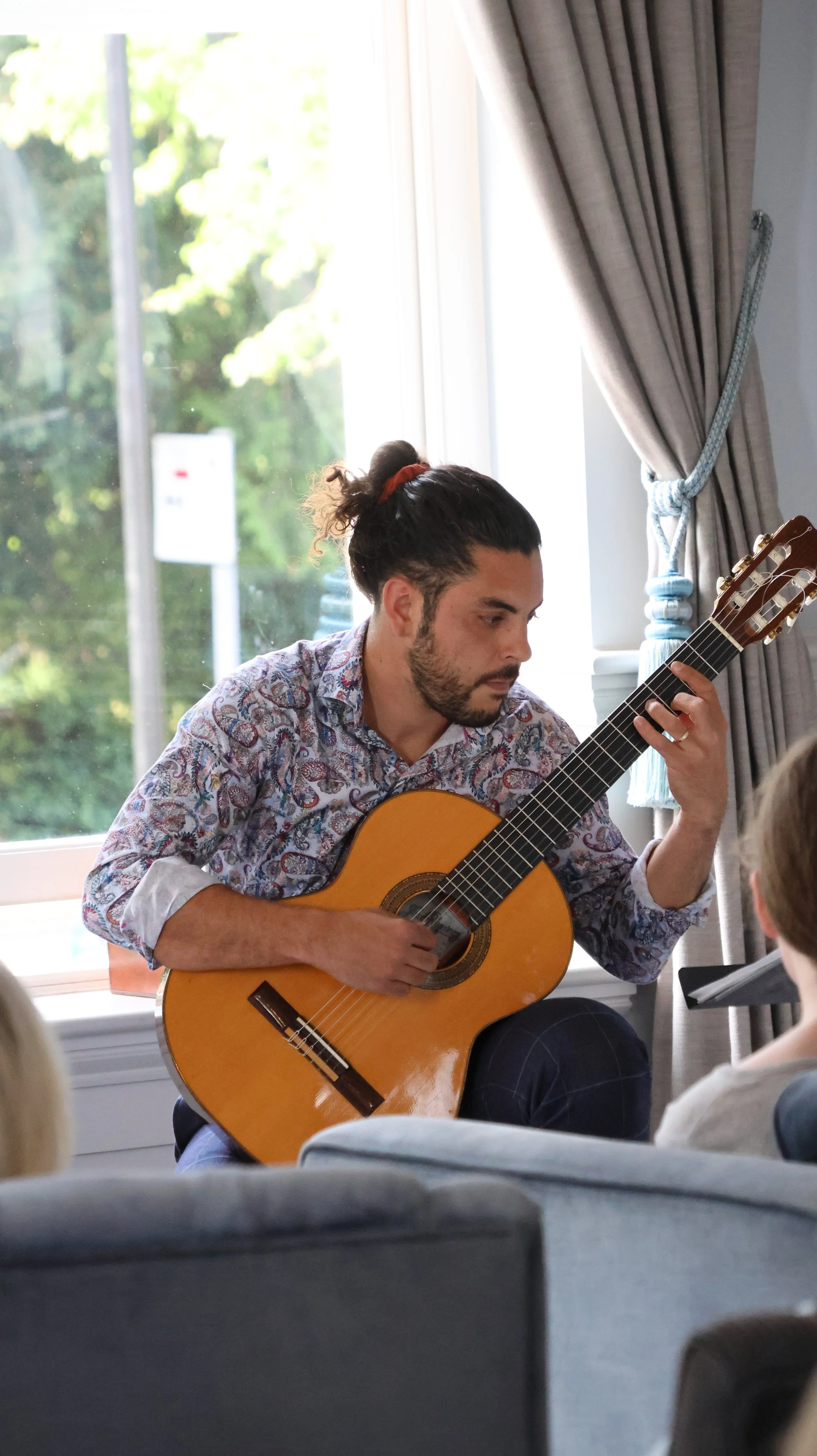 Man playing classical guitar during indoor concert in front of audience, near window with natural light and beige curtains.