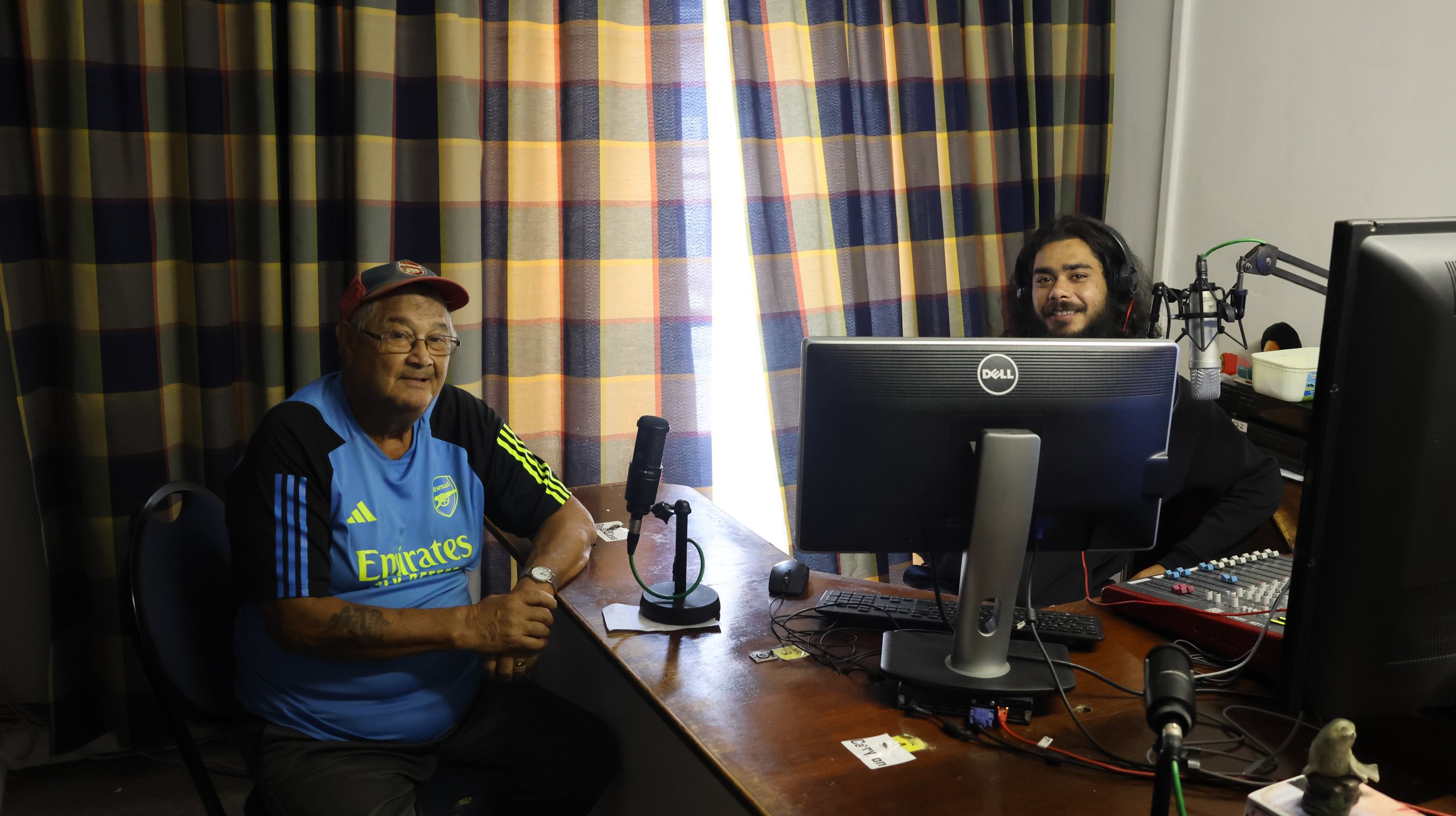 Two men sitting at a radio station, one older wearing a blue sports jersey and cap, the other younger with long hair and headphones, surrounded by radio equipment and a curtain background.