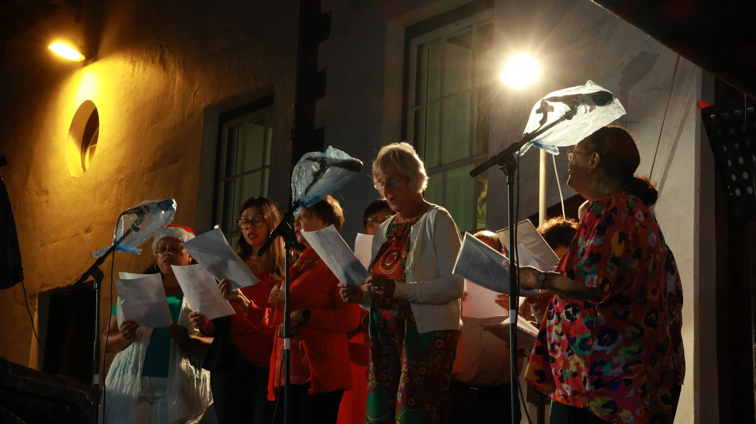 Group of people singing or reading during a Christmas-themed event inside a building, some wearing holiday accessories like Santa hats and colorful clothing, with microphones and sheet music, illuminated by warm lighting.