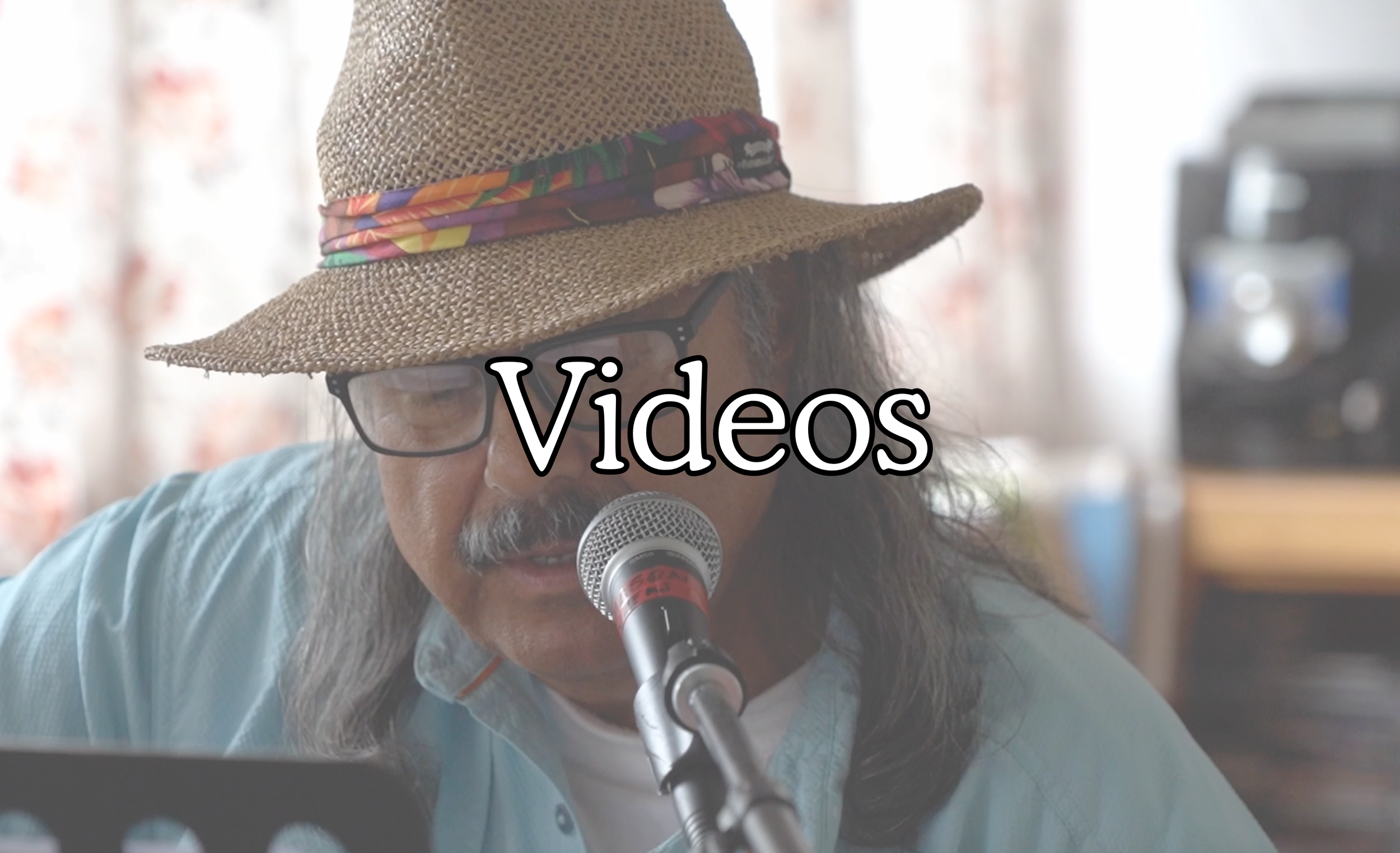 A man wearing glasses and a straw hat with a colorful band is speaking into a microphone. The word 'Videos' is overlaid on the image.