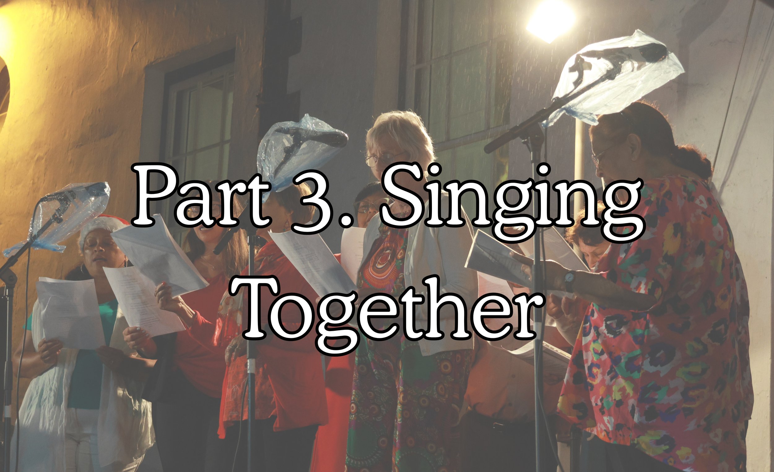 A group of diverse women singing together with sheet music in a room with warm lighting. Some women wear glasses and colorful clothing. There are microphones and a background of a textured wall.