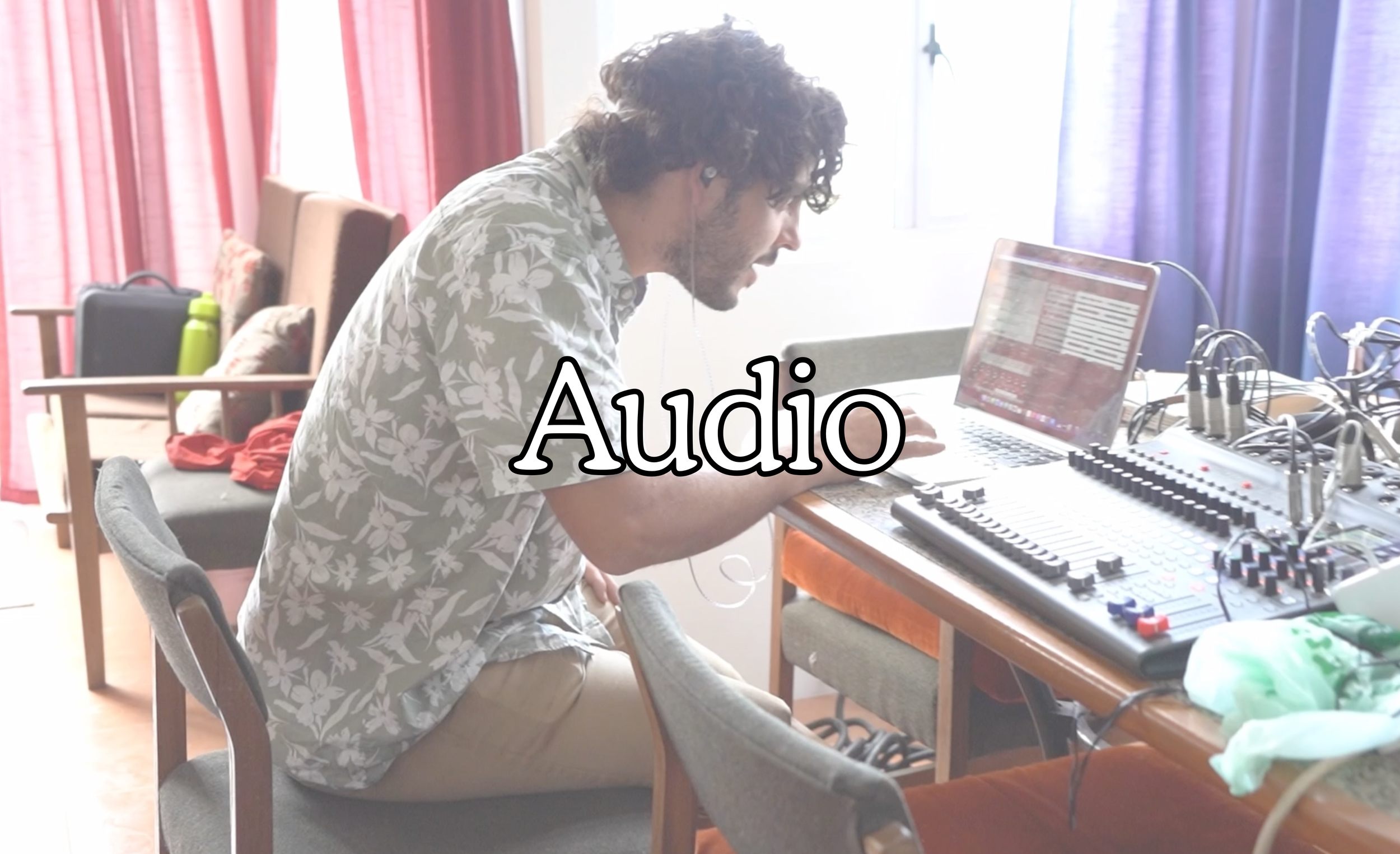 A man with curly hair is sitting at a table with a mixing console, laptop, and headphones, appearing to be working on music production. The room has chairs and curtains with natural light coming from windows.