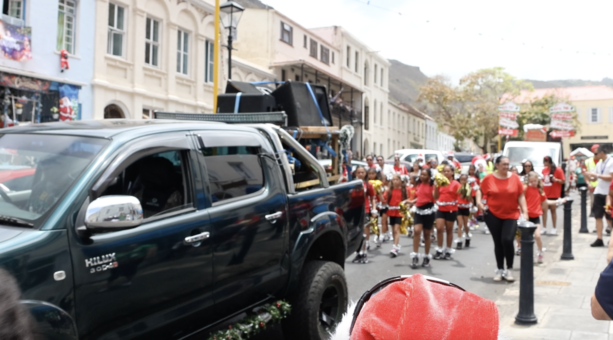 A parade on a city street with a float being pulled by a black pickup truck, and a group of people walking, many dressed in red and some holding cheerleading pom-poms, with decorations and buildings in the background.