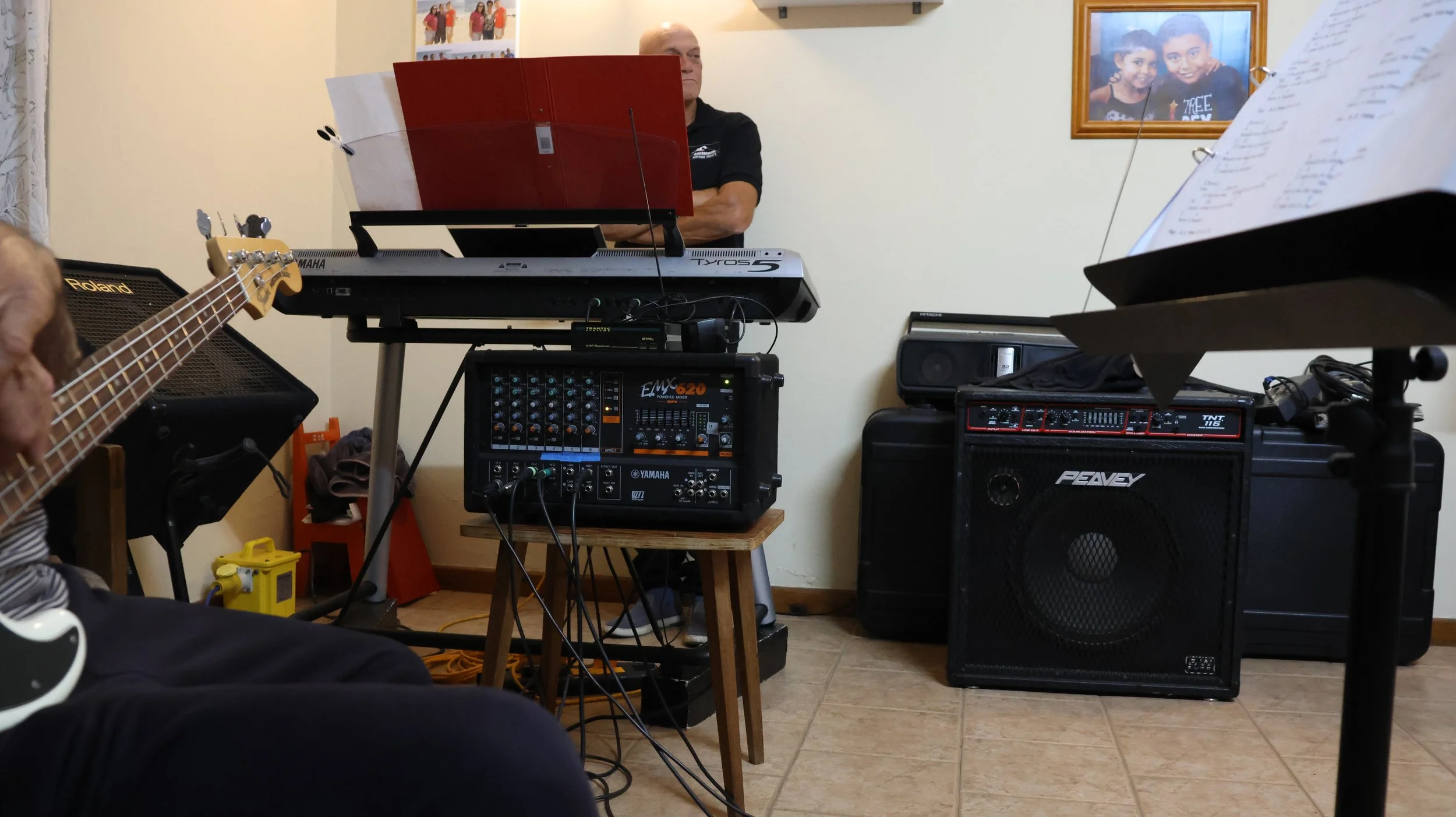 A music rehearsal room with a person playing a bass guitar, a keyboard, and various audio equipment including speakers and a mixer. There is a framed photo of two children hanging on the wall.