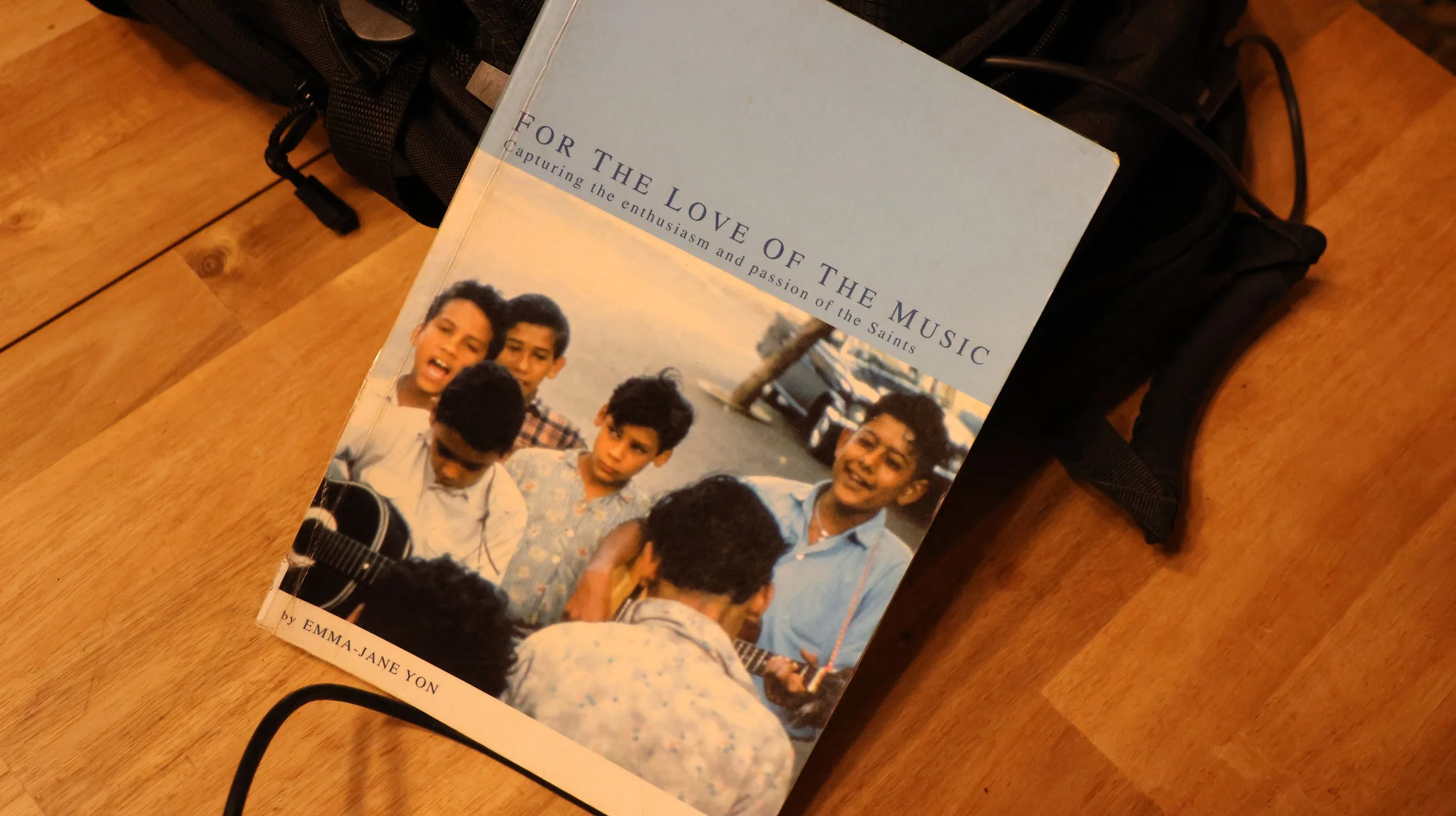 A book titled 'For the Love of the Music' by Emma-Jane Yon resting on a backpack on a wooden floor. The cover features a photo of a group of young men, with some playing a guitar and others singing or looking on.