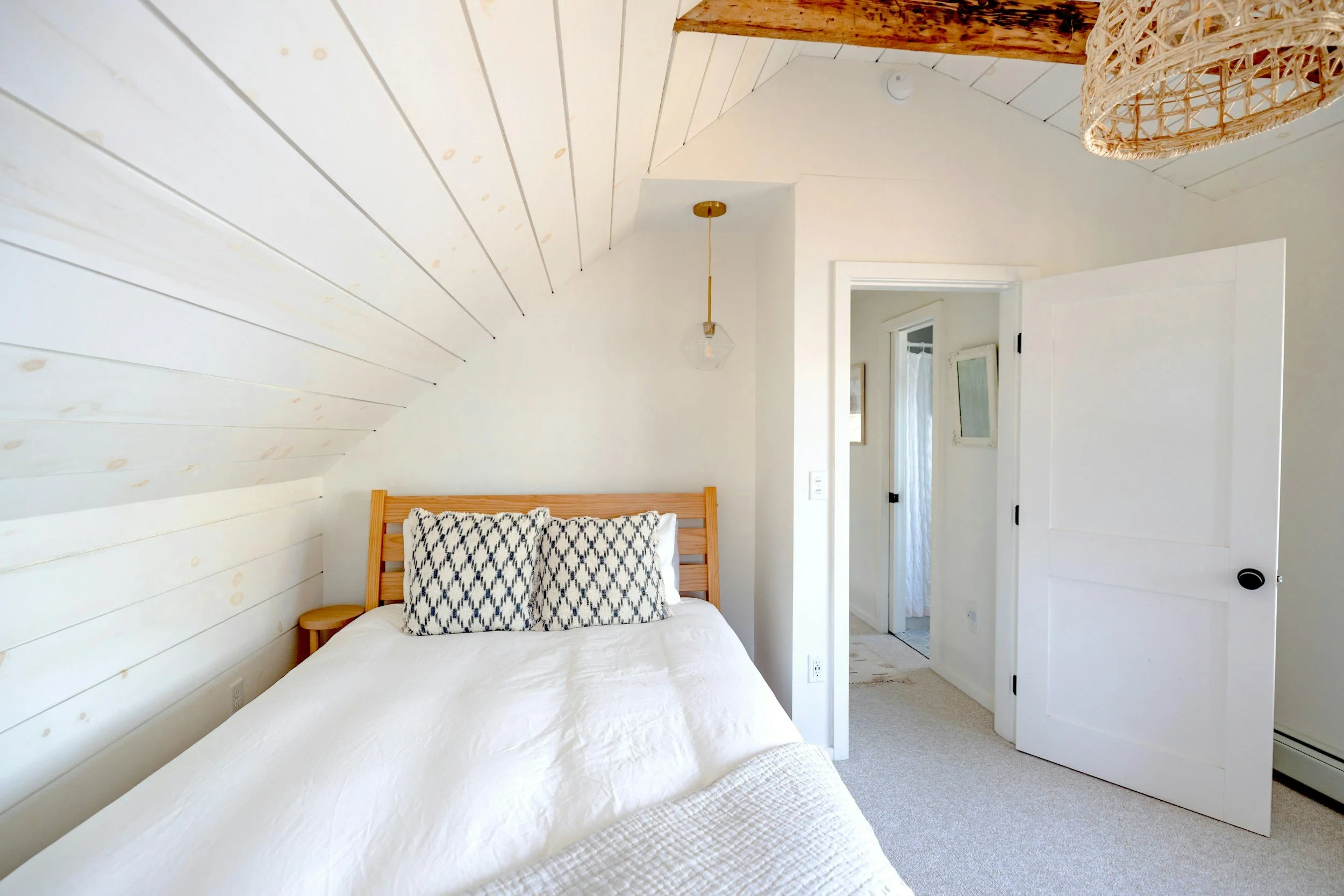 A bedroom with white shiplap walls and ceiling, a bed with white bedding and two black and white patterned pillows, a wooden nightstand, a hanging light fixture, and a partially open white door leading to another room.