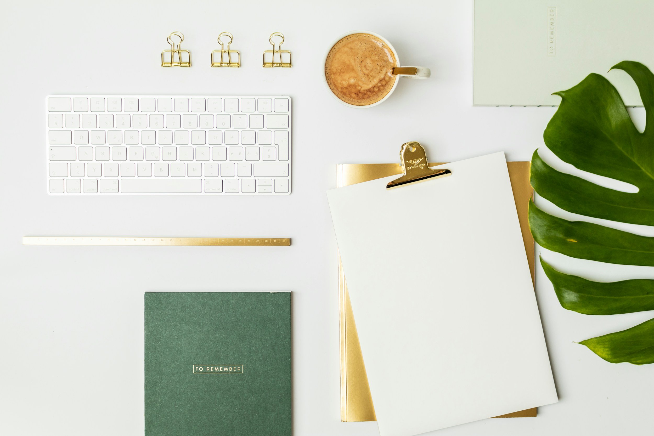 A white desk with a white computer keyboard, three gold binder clips, a cup of coffee, a closed light green notebook, a gold clipboard with a blank white sheet, a gold ruler, and a dark green notebook labeled "TO REMEMBER." There is a large green plant leaf on the right side of the desk.