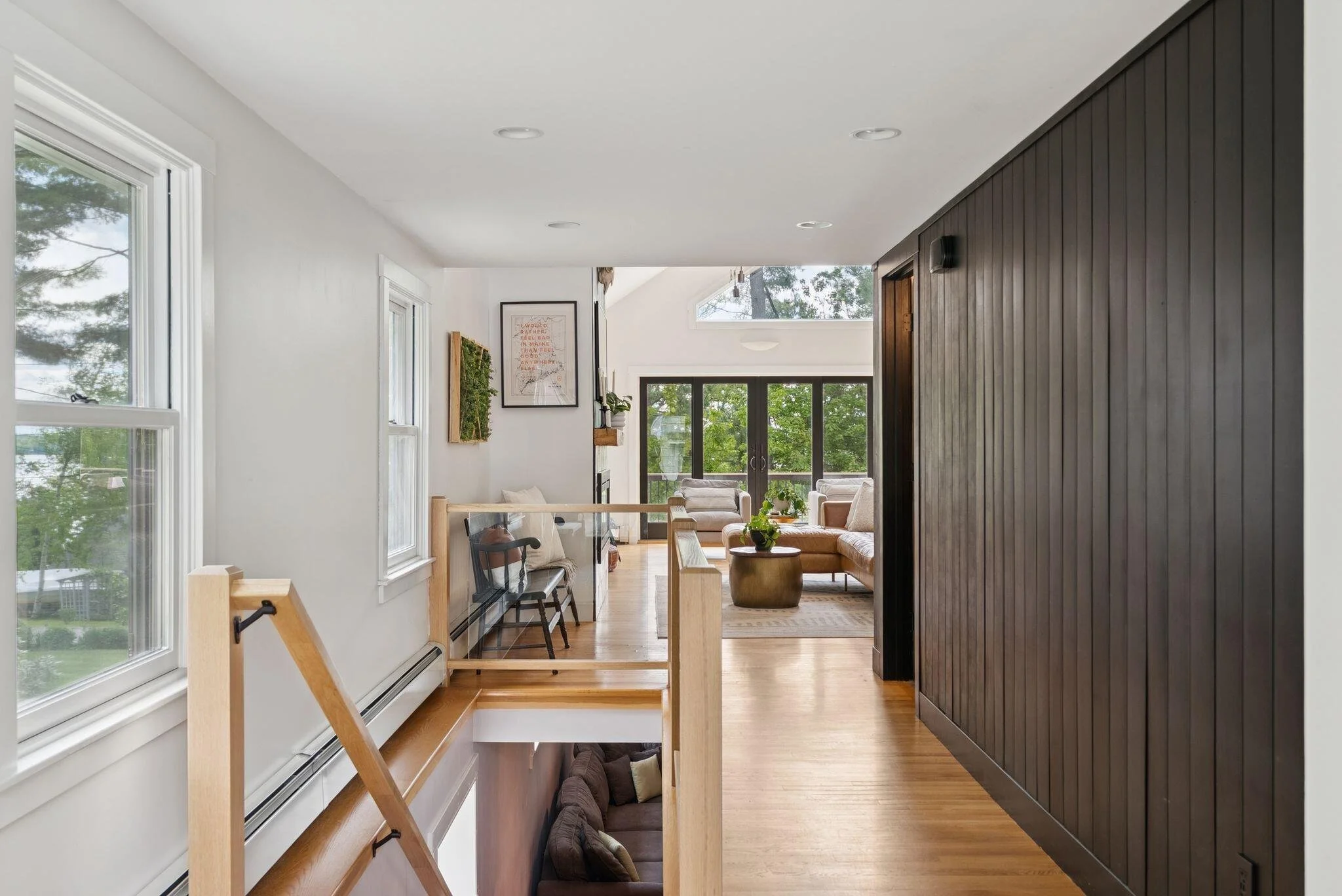A bright living room with large windows, white walls, wooden floors, and modern furniture including armchairs and a coffee table. There is a dark wood paneled wall on the right and various decorative elements on the walls, with a view of greenery outside.