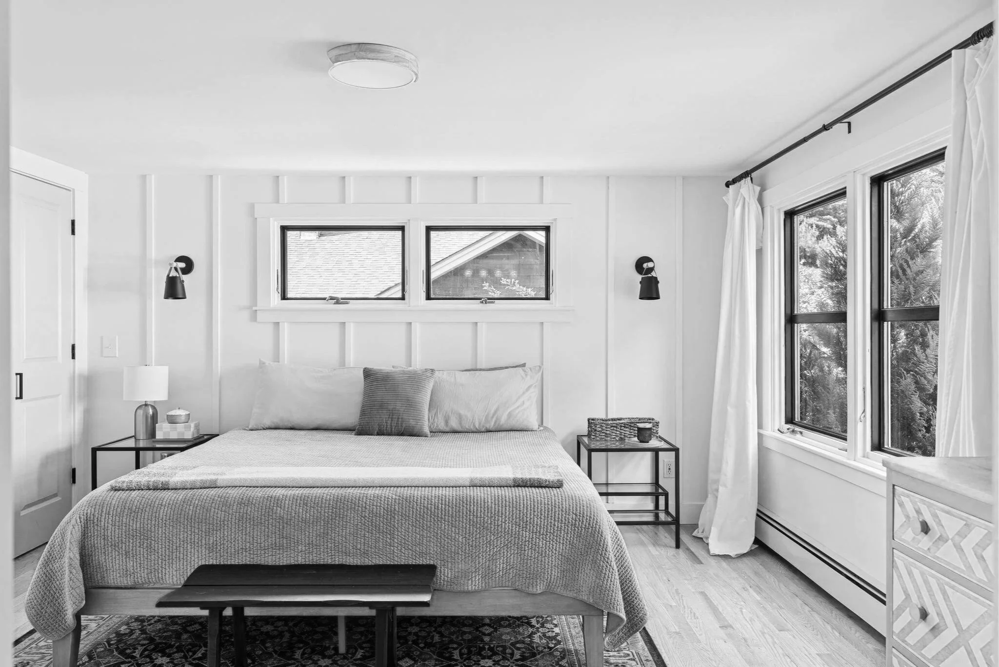 A bedroom with light-colored walls and large windows with white curtains, a bed with a textured bedspread and pillows, and black wall-mounted lamps on either side of the window.