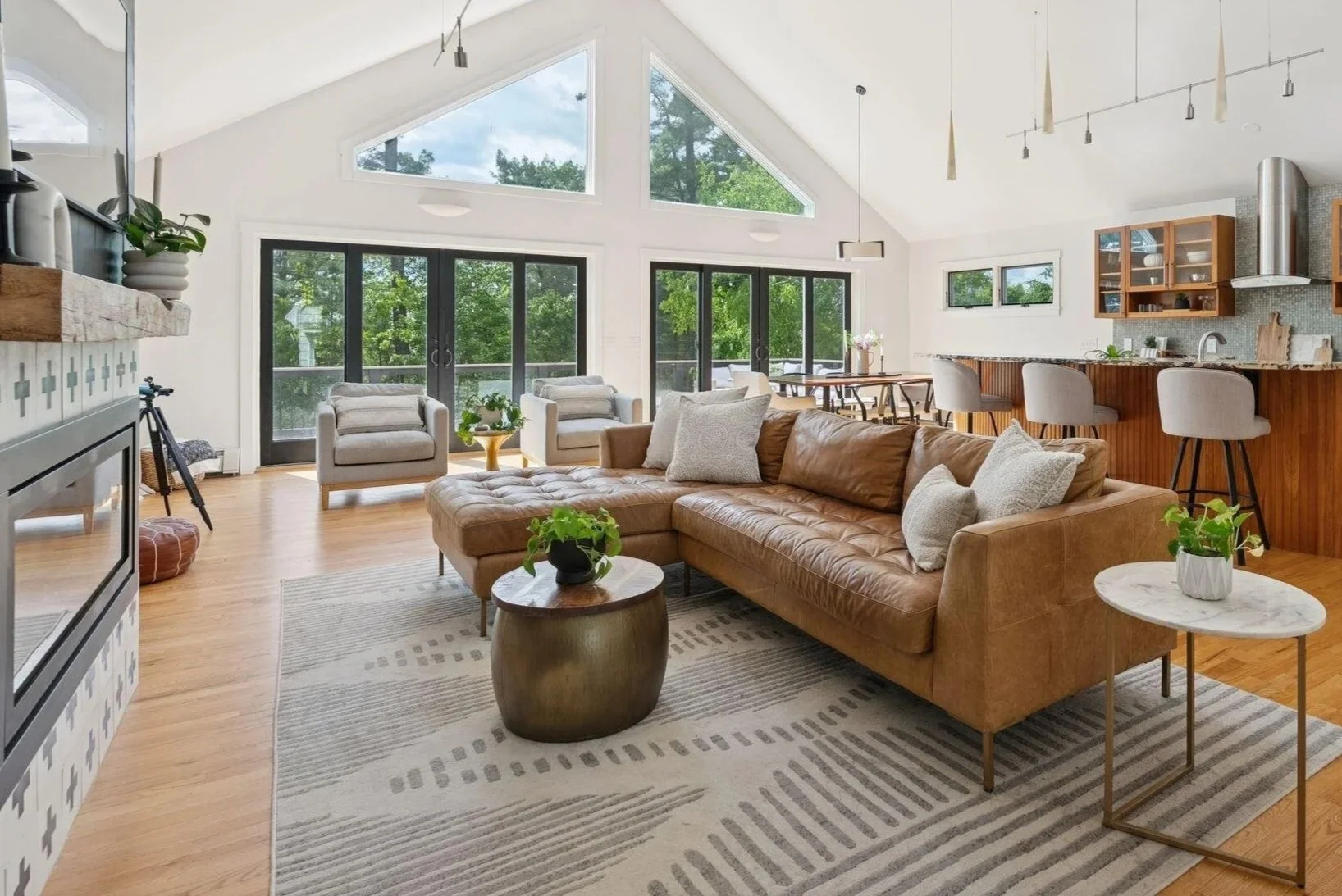 Open-concept living room and kitchen with large windows, tan leather sectional, white chairs, and wooden accents.