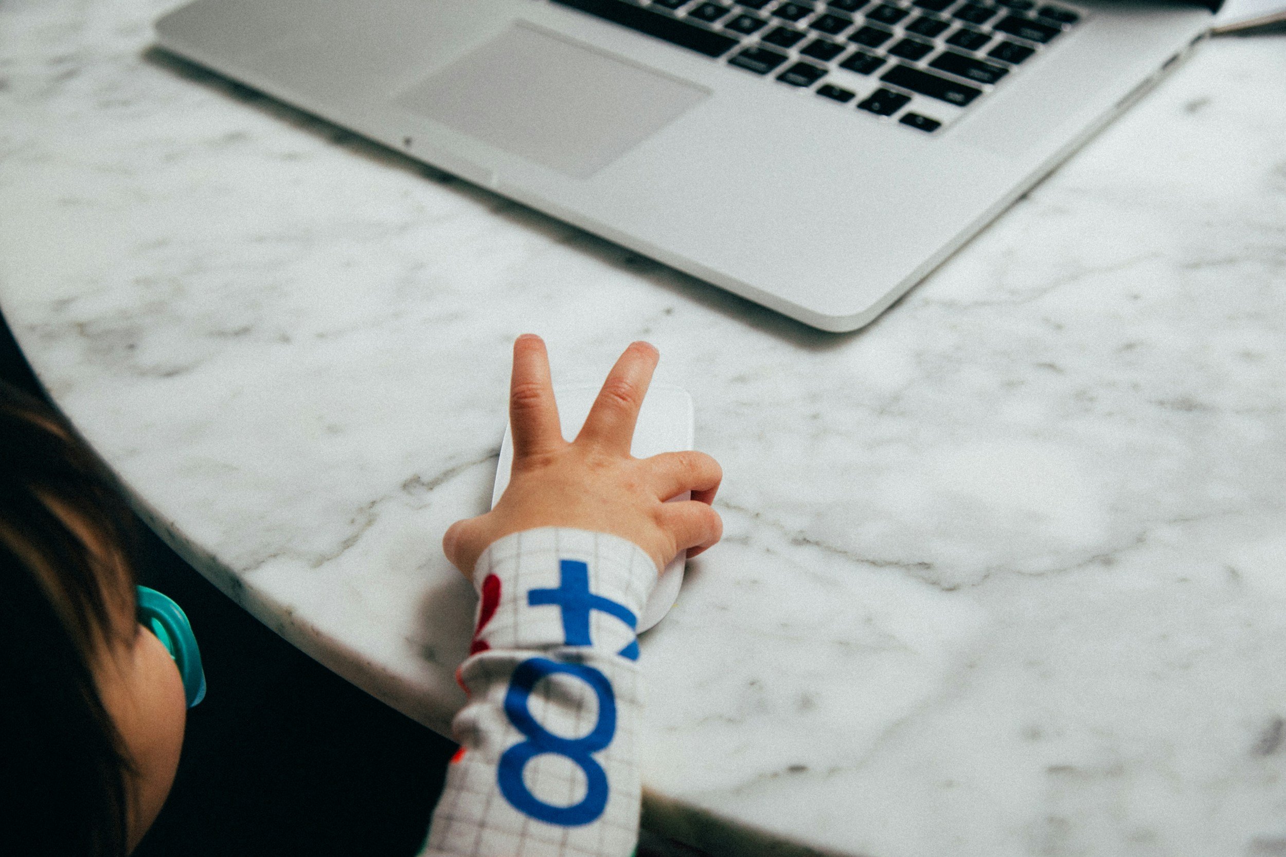 Child's hand making a peace sign near a laptop on a marble surface.