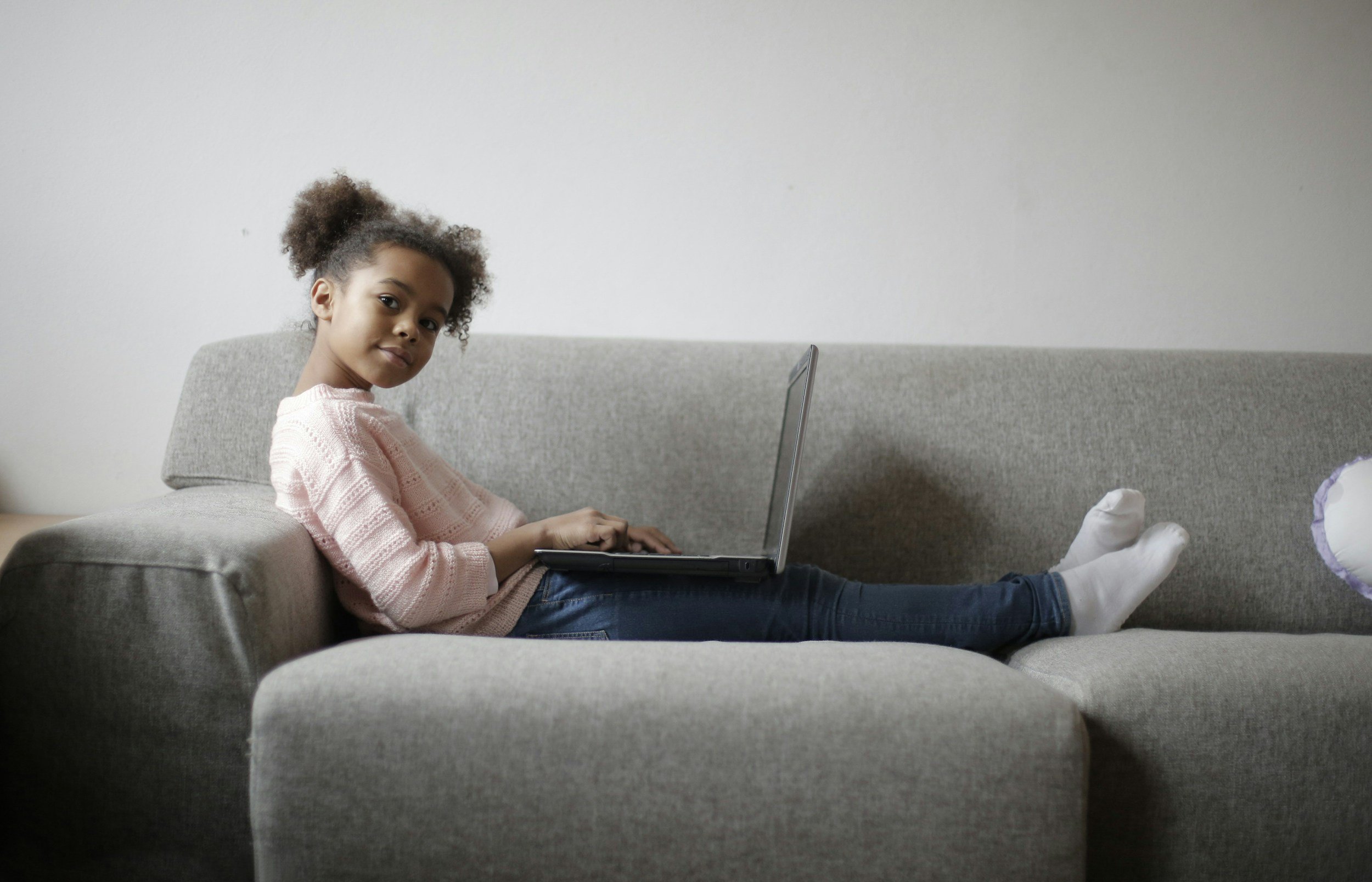 A young girl with curly hair sitting on a gray couch, using a laptop, wearing a pink sweater and blue jeans with white socks, in a room with plain white walls.
