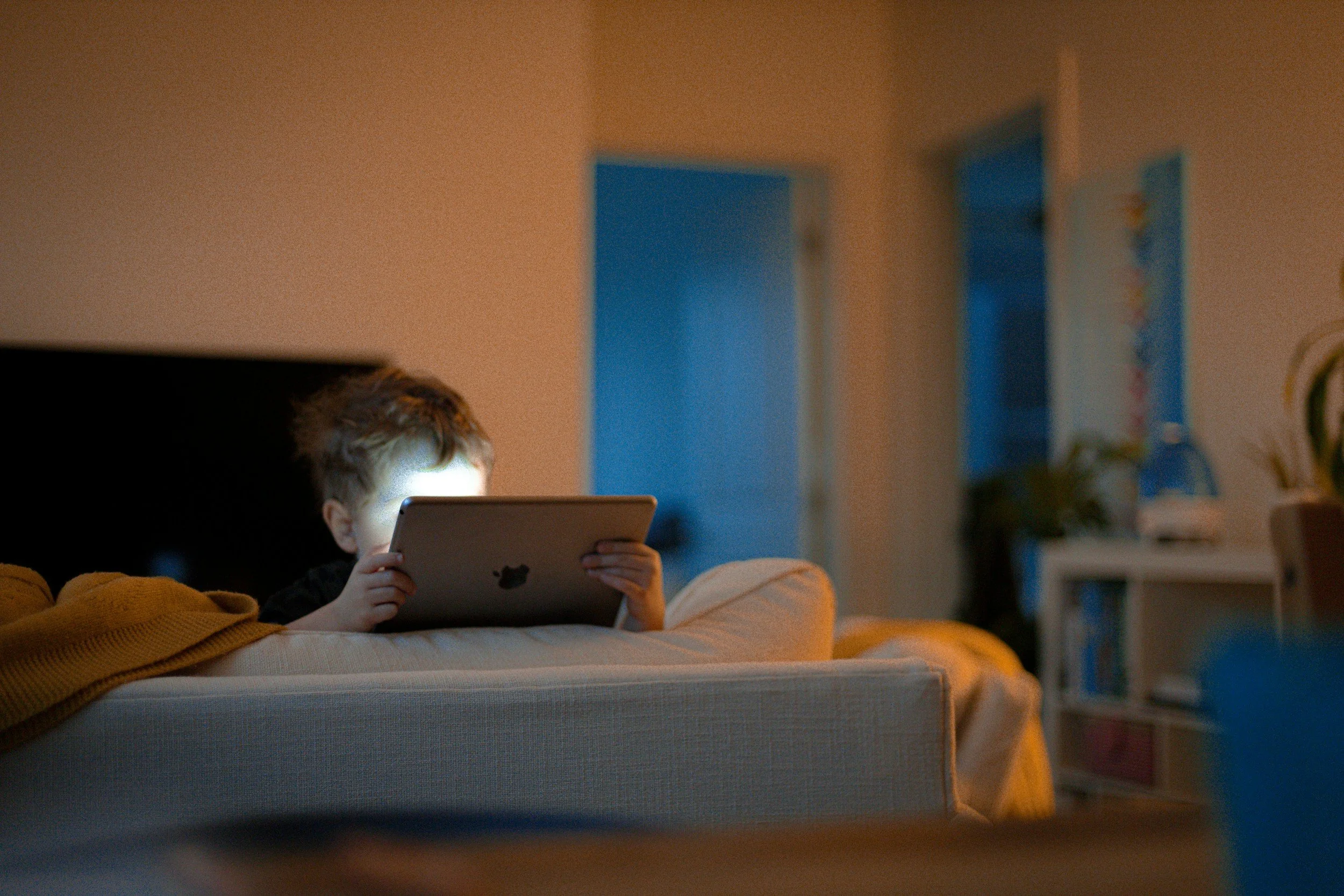 A young boy sitting on a couch at night, illuminated by a tablet screen, with a blurred living room background, including a doorway and a bookshelf.