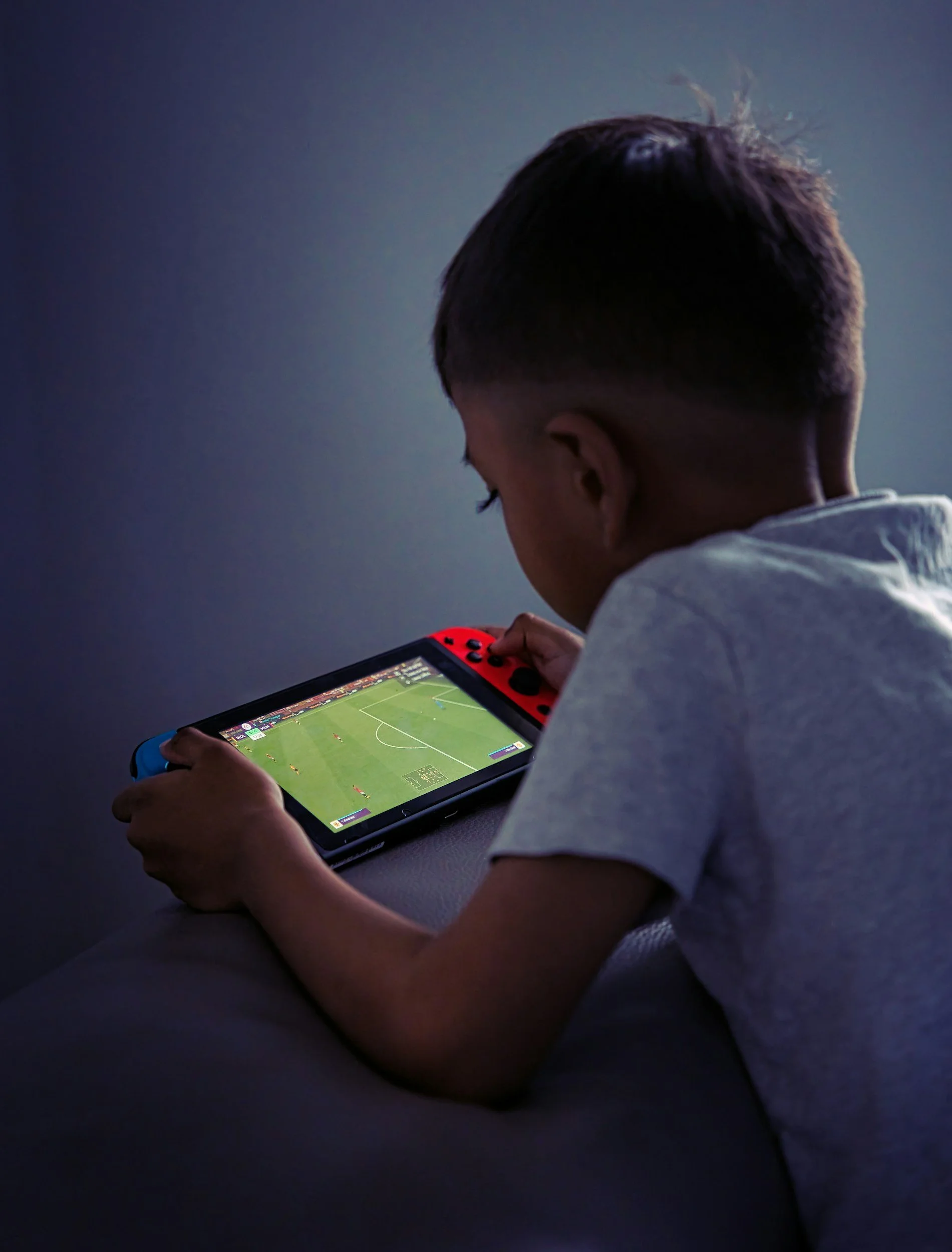 A young person playing a soccer video game on a handheld gaming device in a dimly lit room.