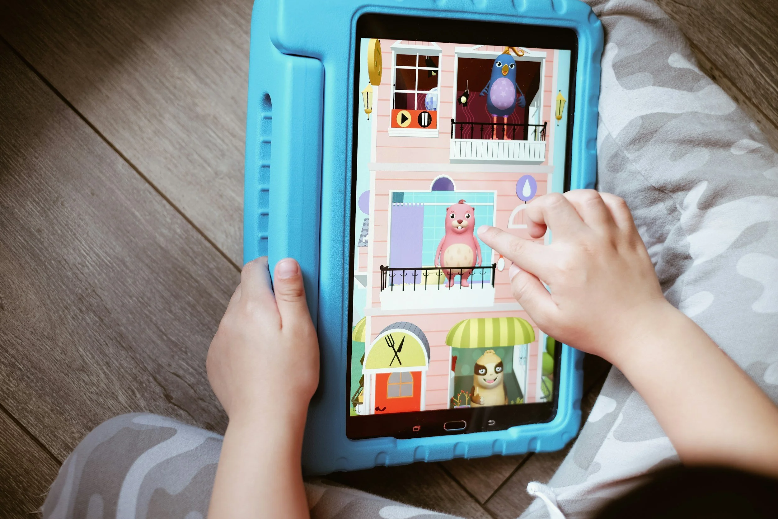 Children playing a game on a tablet with cartoon characters on the screen, sitting on a wooden floor.