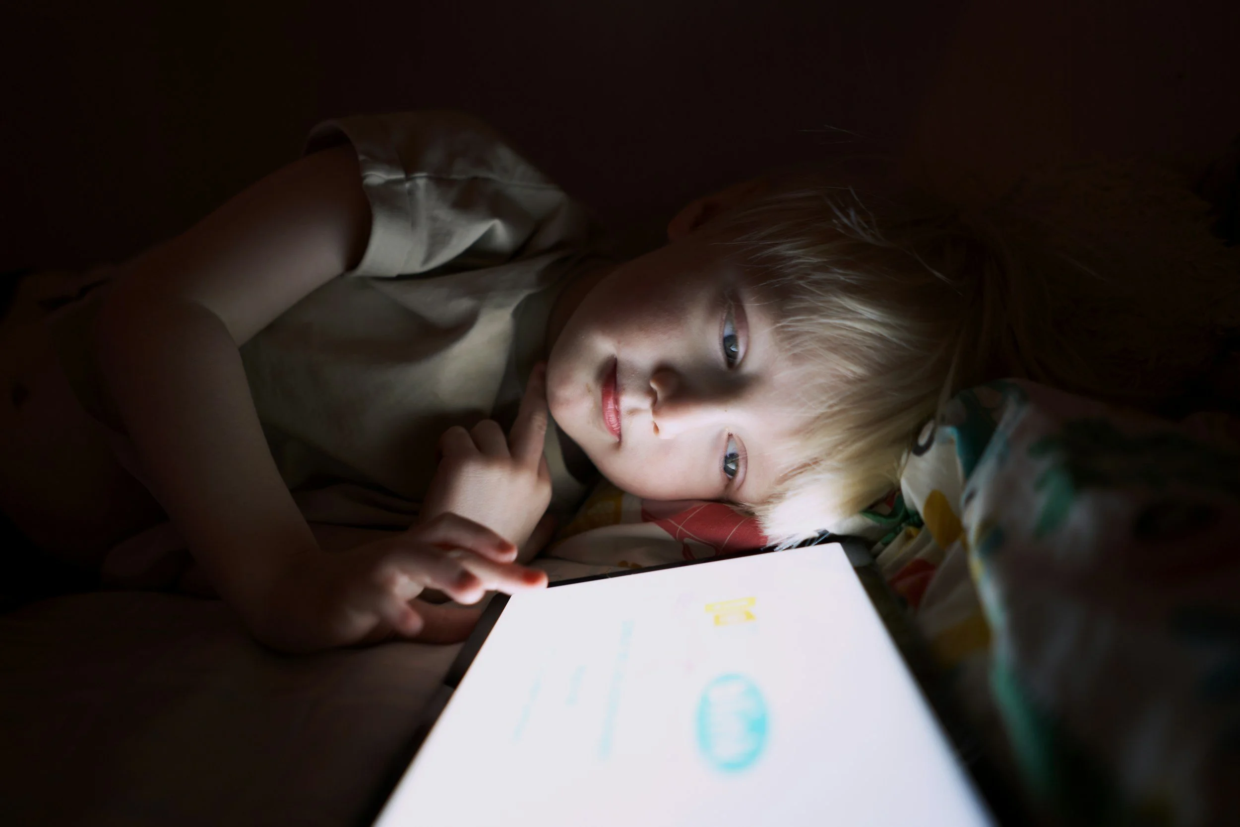 A young child with blonde hair looking at a tablet screen in a dark room, with the screen illuminating their face.