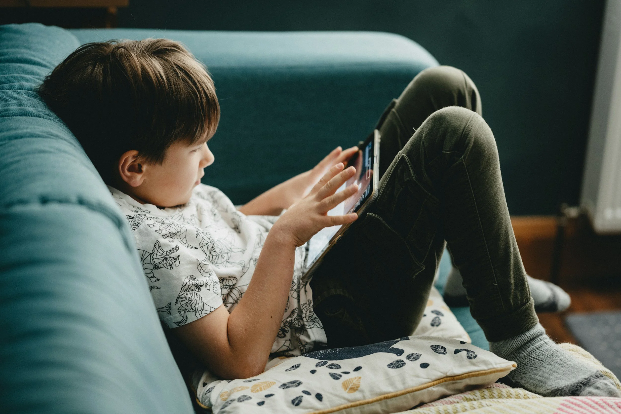 A young boy with brown hair is lying on a teal couch, focused on a smartphone in his hands. He is wearing a white shirt with black animals printed on it, dark green pants, and gray socks. The boy is sitting with his legs bent and resting on the couch, with a decorative throw blanket with leaf patterns partially visible in front of him.