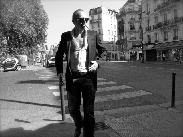 A man in a suit with sunglasses standing on a city street crossing, with buildings and cars in the background.