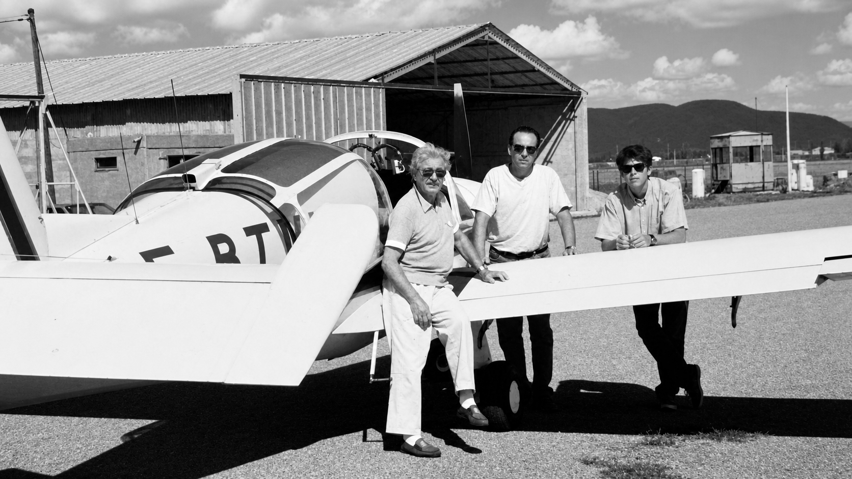 Three men stand next to a small airplane outdoors in front of a building with mountains in the background, in black and white.