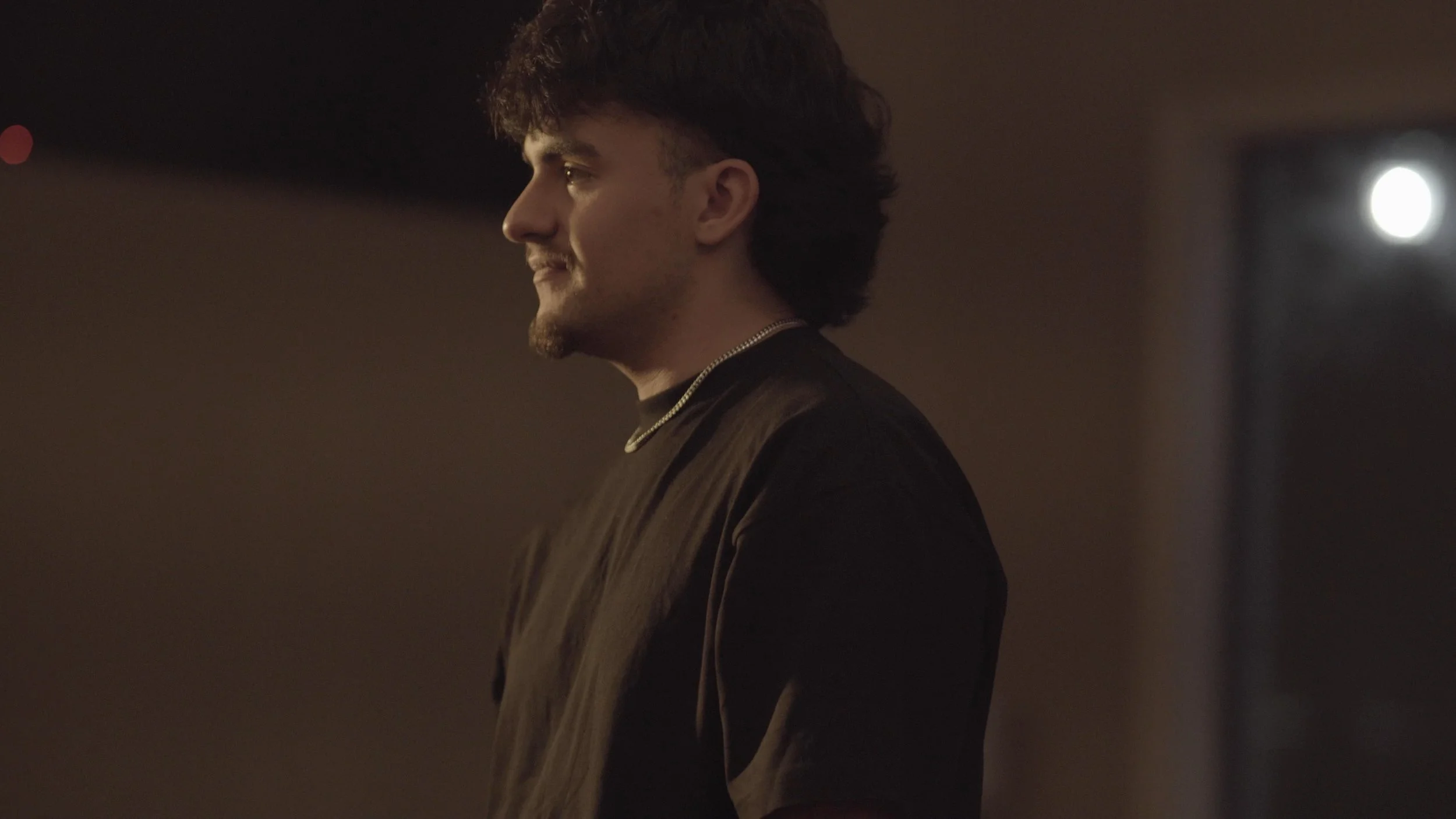 Side profile of a young man with dark, curly hair and a light beard, wearing a dark shirt and a silver chain, standing in a dimly lit room with a blurred bright light in the background.