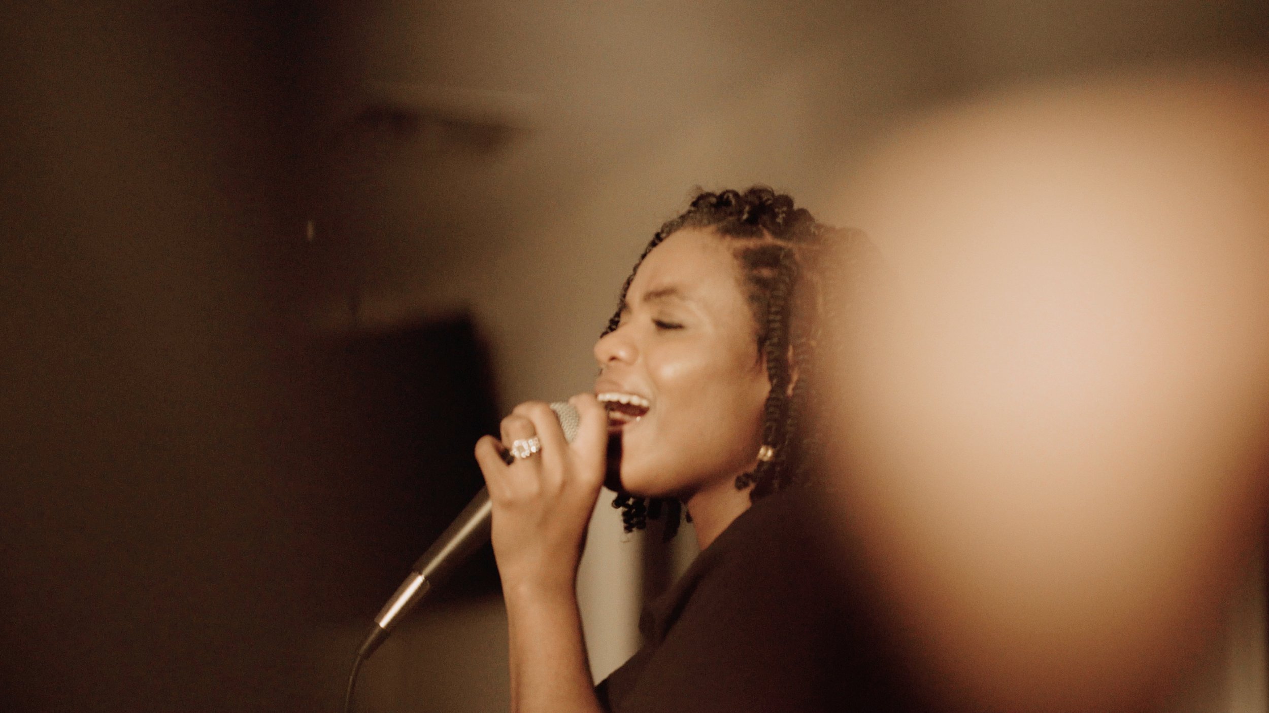 A woman with braided hair singing into a microphone, eyes closed and wearing a black outfit.