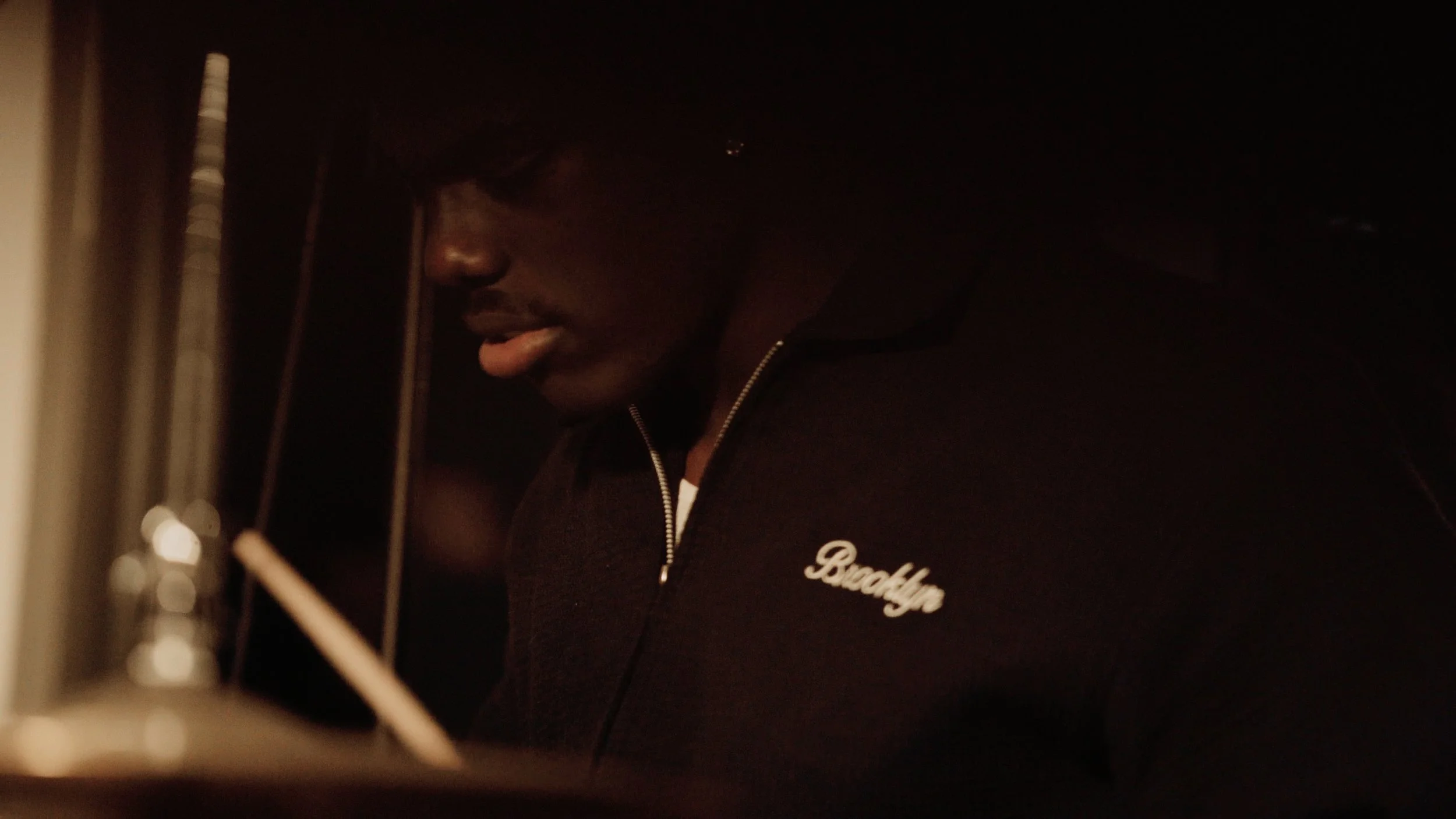 A person with dark skin wearing glasses and a black hoodie that says 'Brooklyn' is in a recording studio.