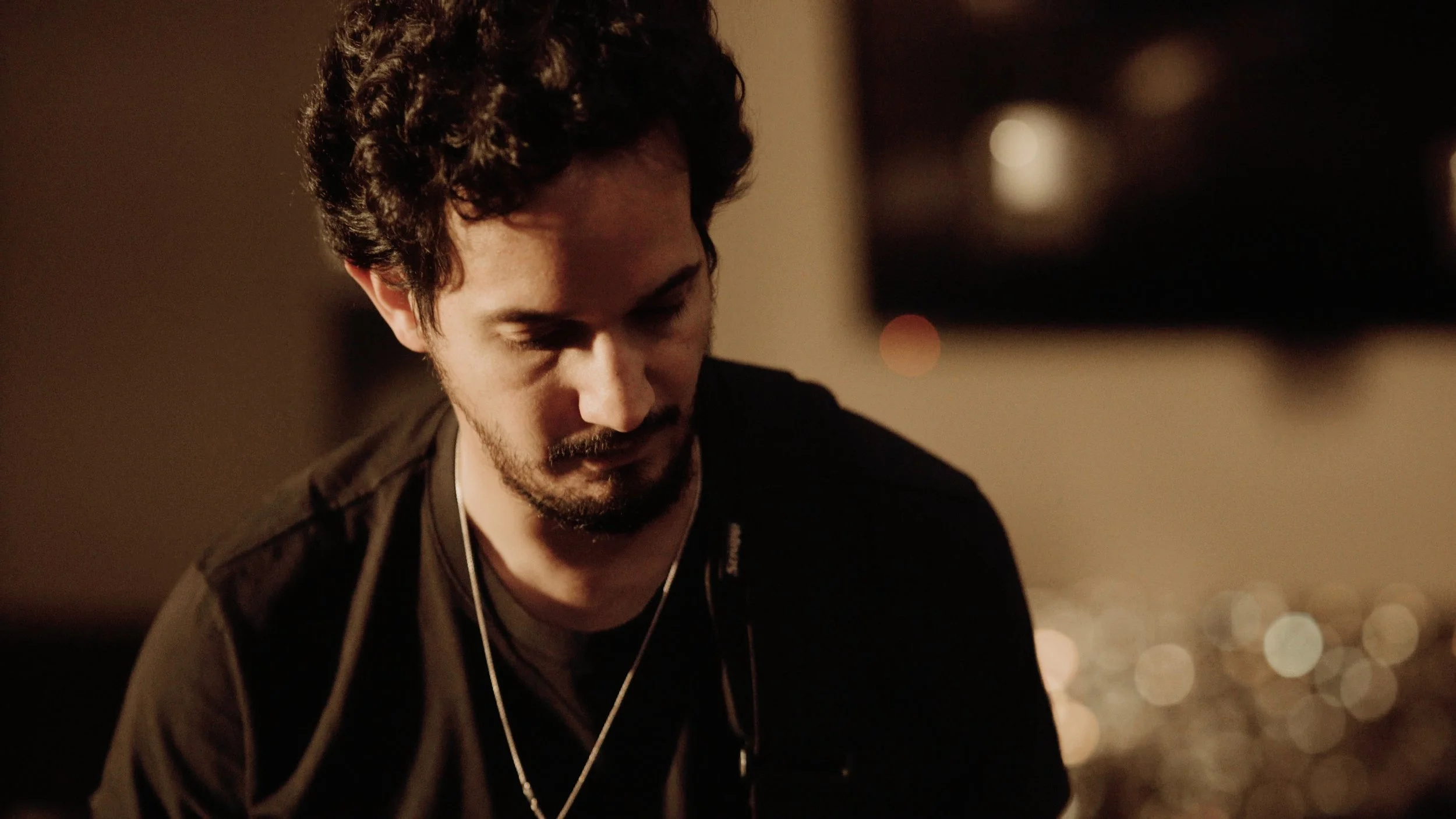 A young man with curly dark hair and a beard wearing headphones, looking down thoughtfully in a dimly lit room with blurred city lights in the background.