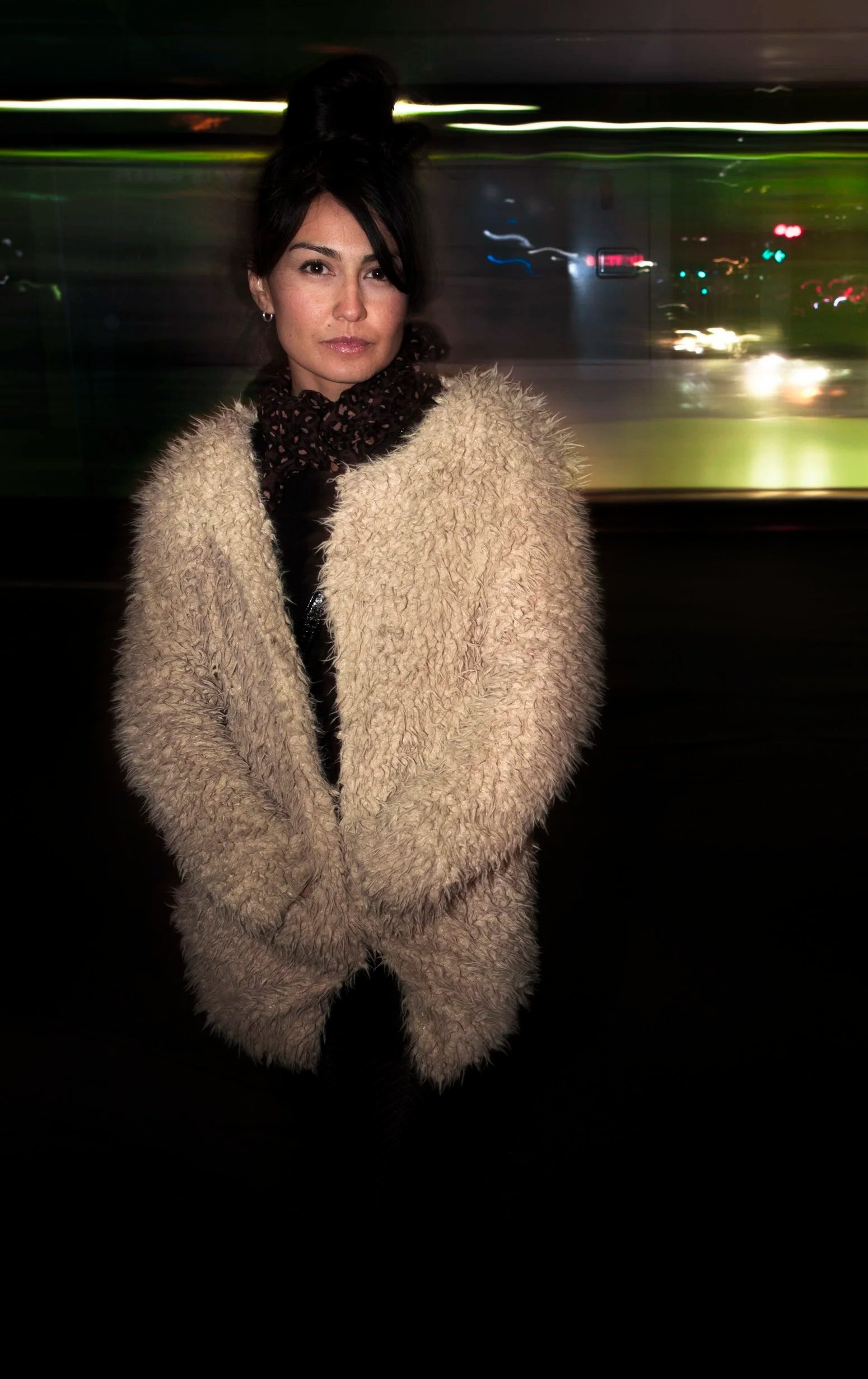 A woman with dark hair styled in a high bun, wearing a beige fuzzy coat and a dark leopard-print scarf, standing in front of a blurred cityscape at night.
