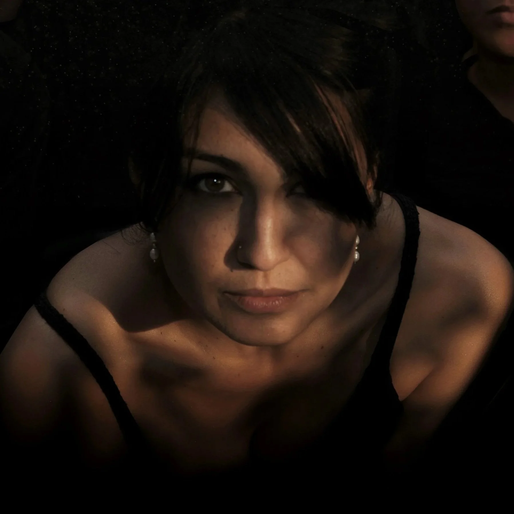 Close-up photo of a woman with dark hair styled with a side-swept fringe, wearing pearl earrings and black attire, with her face partially visible in a low-light setting.