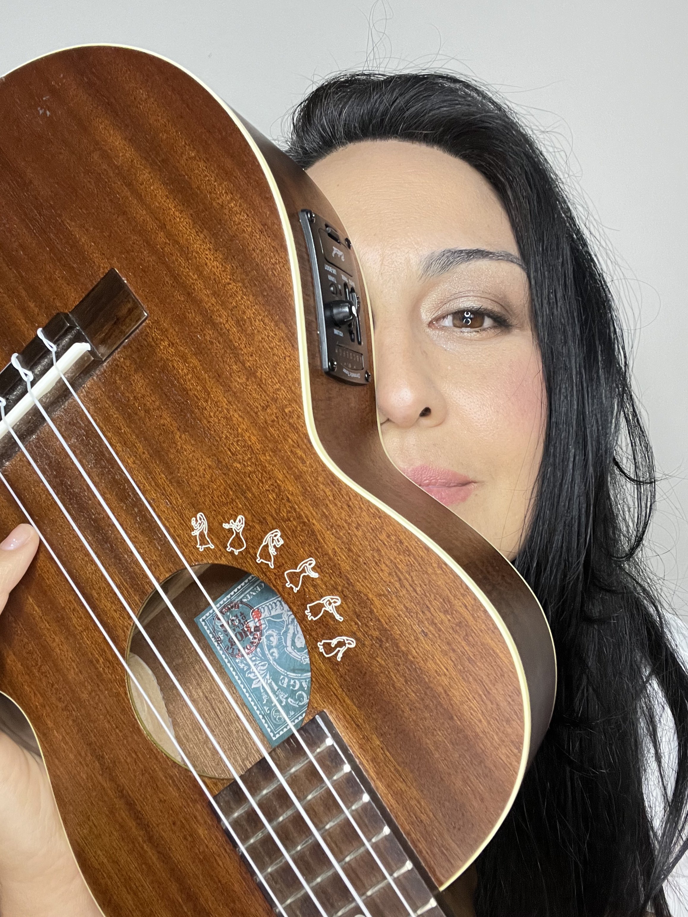 A woman with black hair partially hidden behind an acoustic guitar with a wooden finish, focusing on her eye and part of her face.