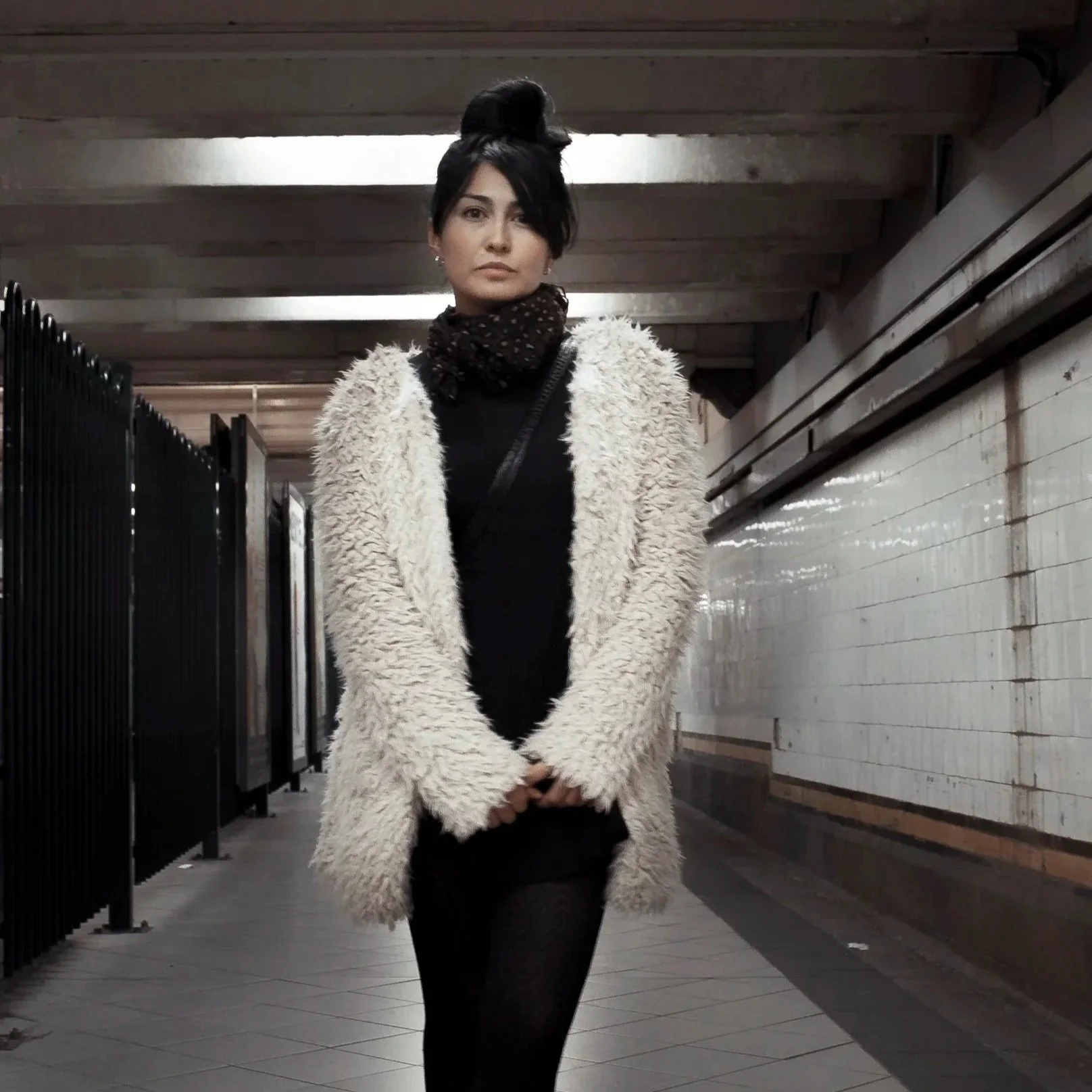 A woman standing in an underground subway station, wearing a fluffy white coat, a black top, black pants, and a dark scarf, with her dark hair styled in a high updo.