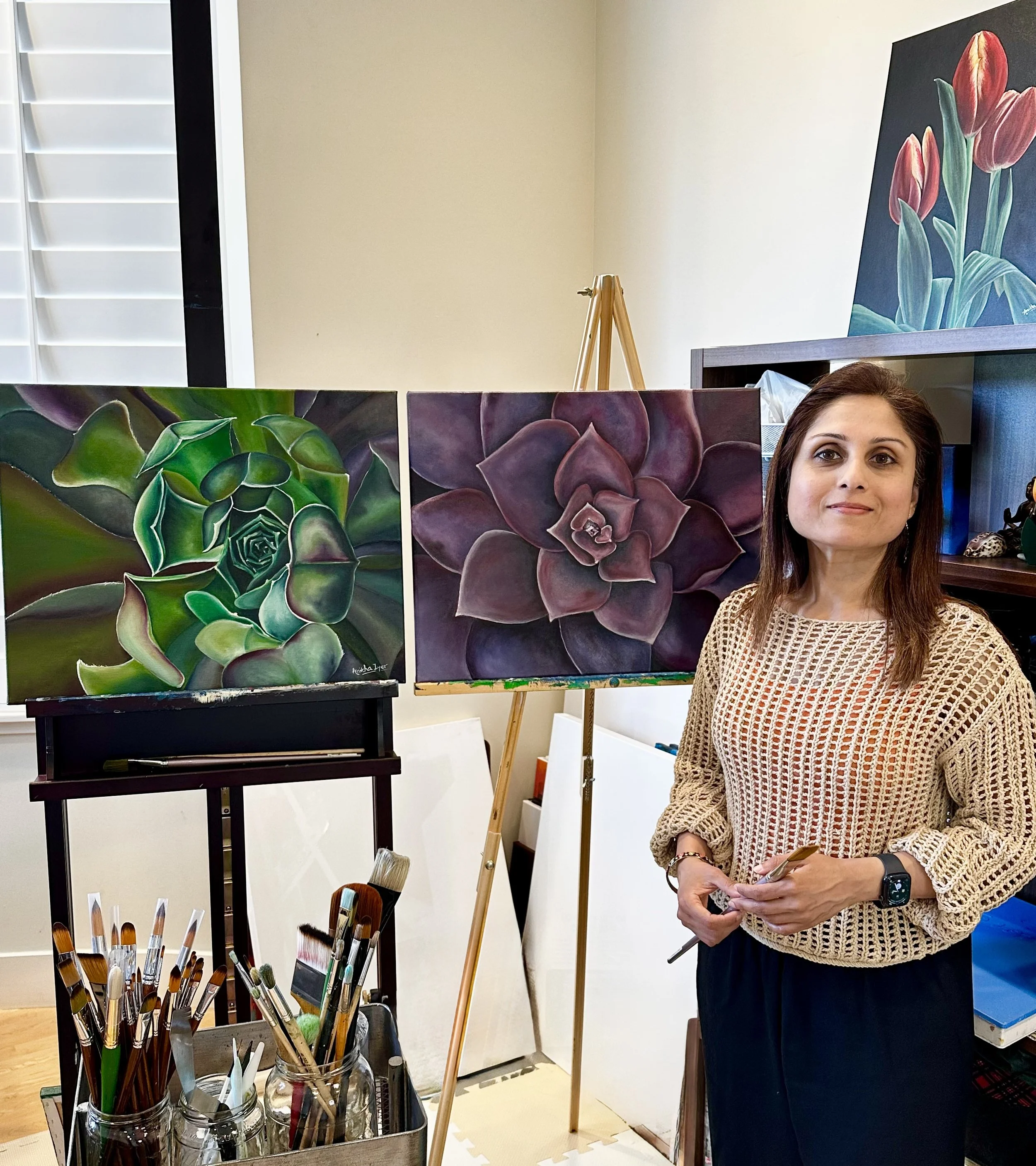 Anitha Iyer art studio. A woman standing in an art studio surrounded by paintings of succulents and tulips. Art supplies, including brushes in mason jars, are in the foreground.