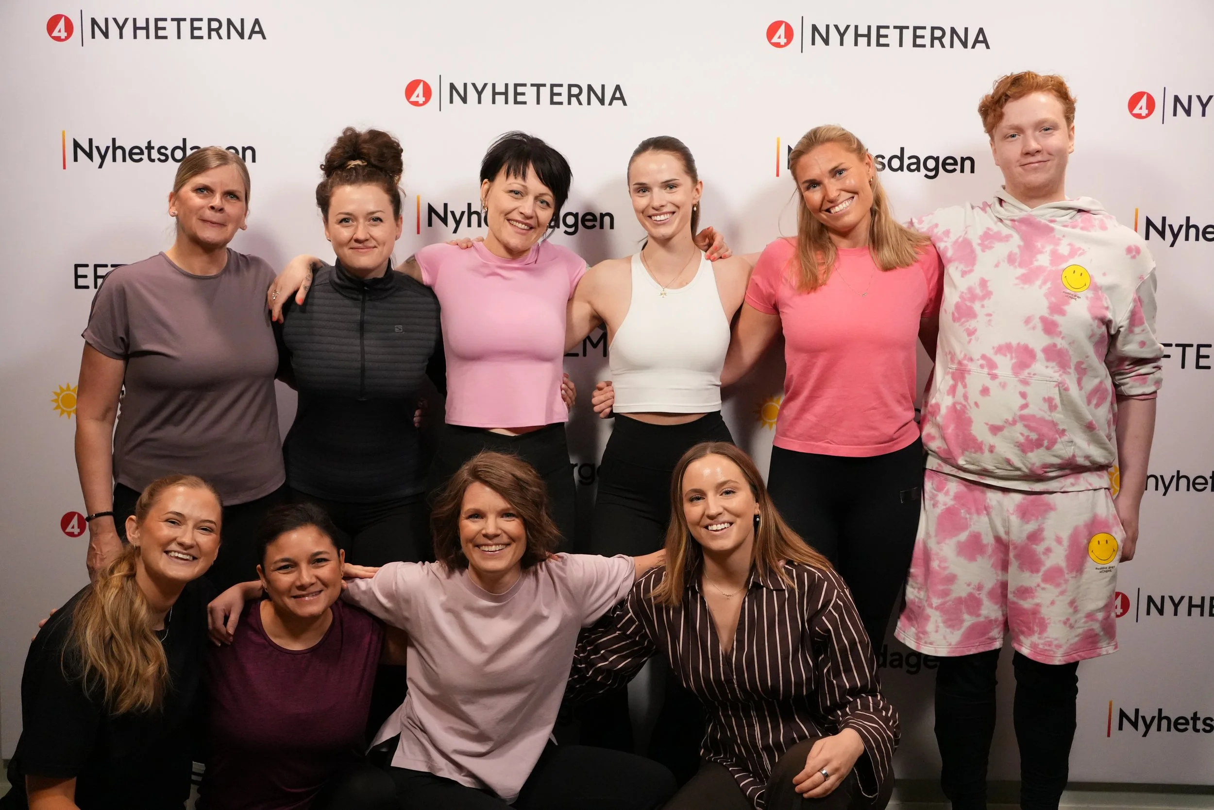 En grupp människor poserar för en gruppbild framför en bakgrund med text och logotyper. De ler och har armar runt varandra, klädda i vardagskläder. Bakgrunden har texten 'NYHETERNA', 'Nyhetsdagen', och logos för '4' och 'NY'.