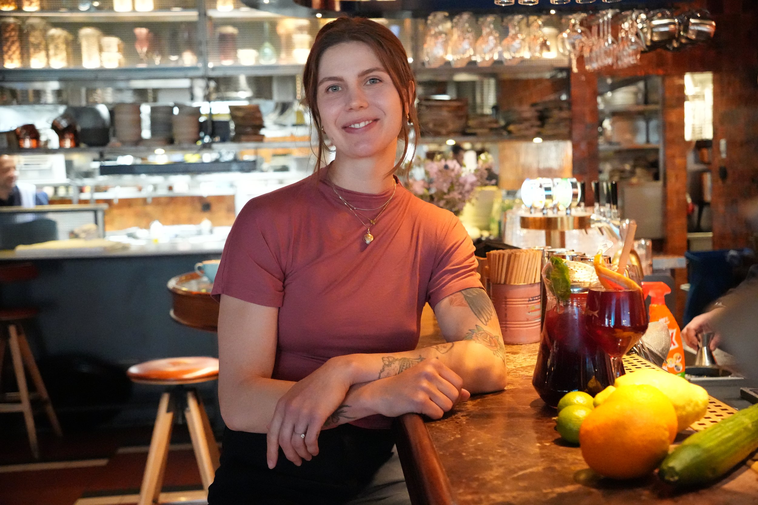 En ung kvinna med tatuering på armen, brunt hår, sitter vid ett barbord i en restaurang eller bar, omgiven av färska citrusfrukter. Hon ler och tittar mot kameran.