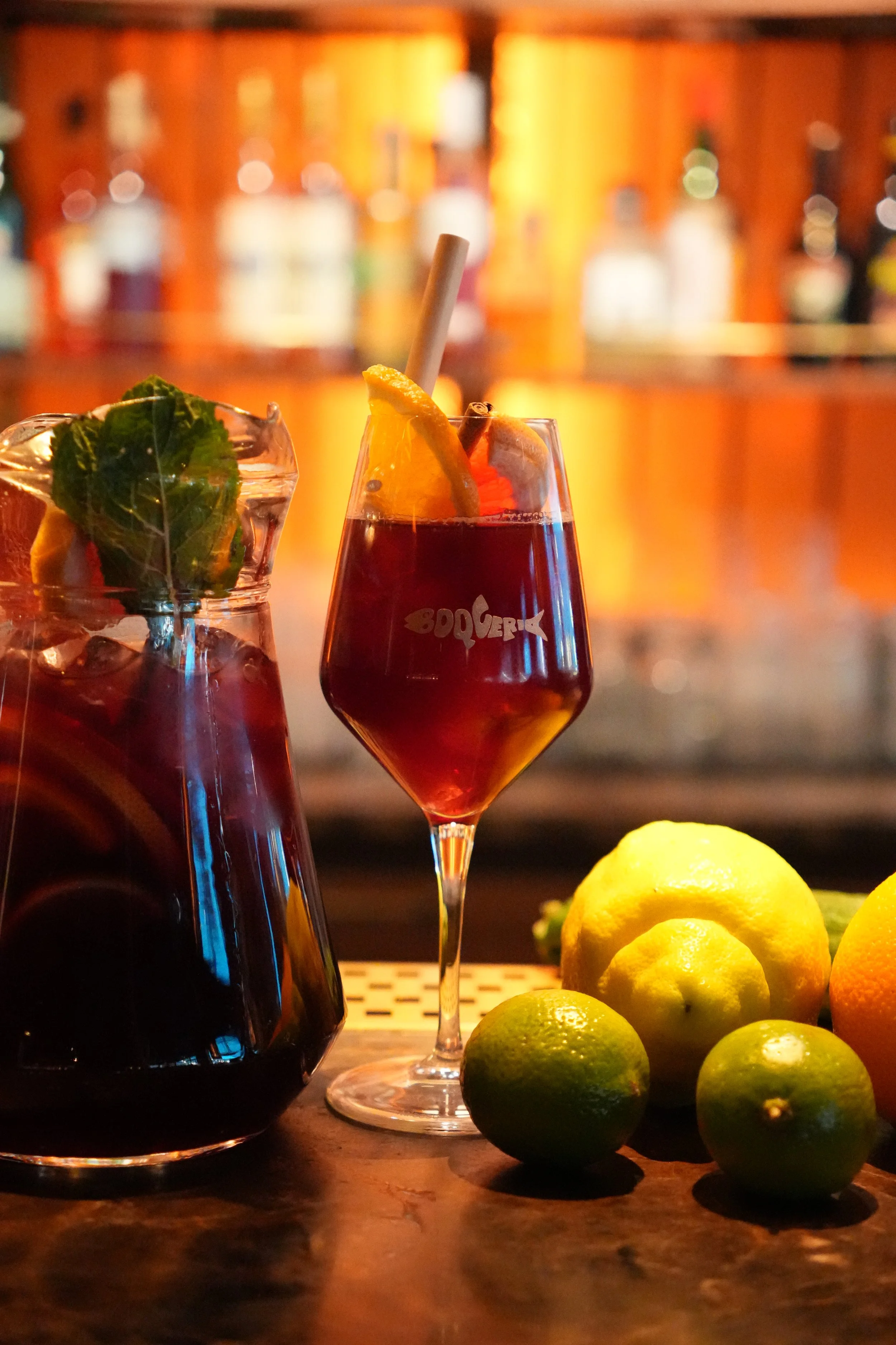 En mörk cocktail i ett cocktailglas, dekorerad med en citronskiva och kanelstång. En annan drink i ett glas med grönt blad och citron skivor, samt flera citroner och lime på bordet. En barhimmel i bakgrunden.